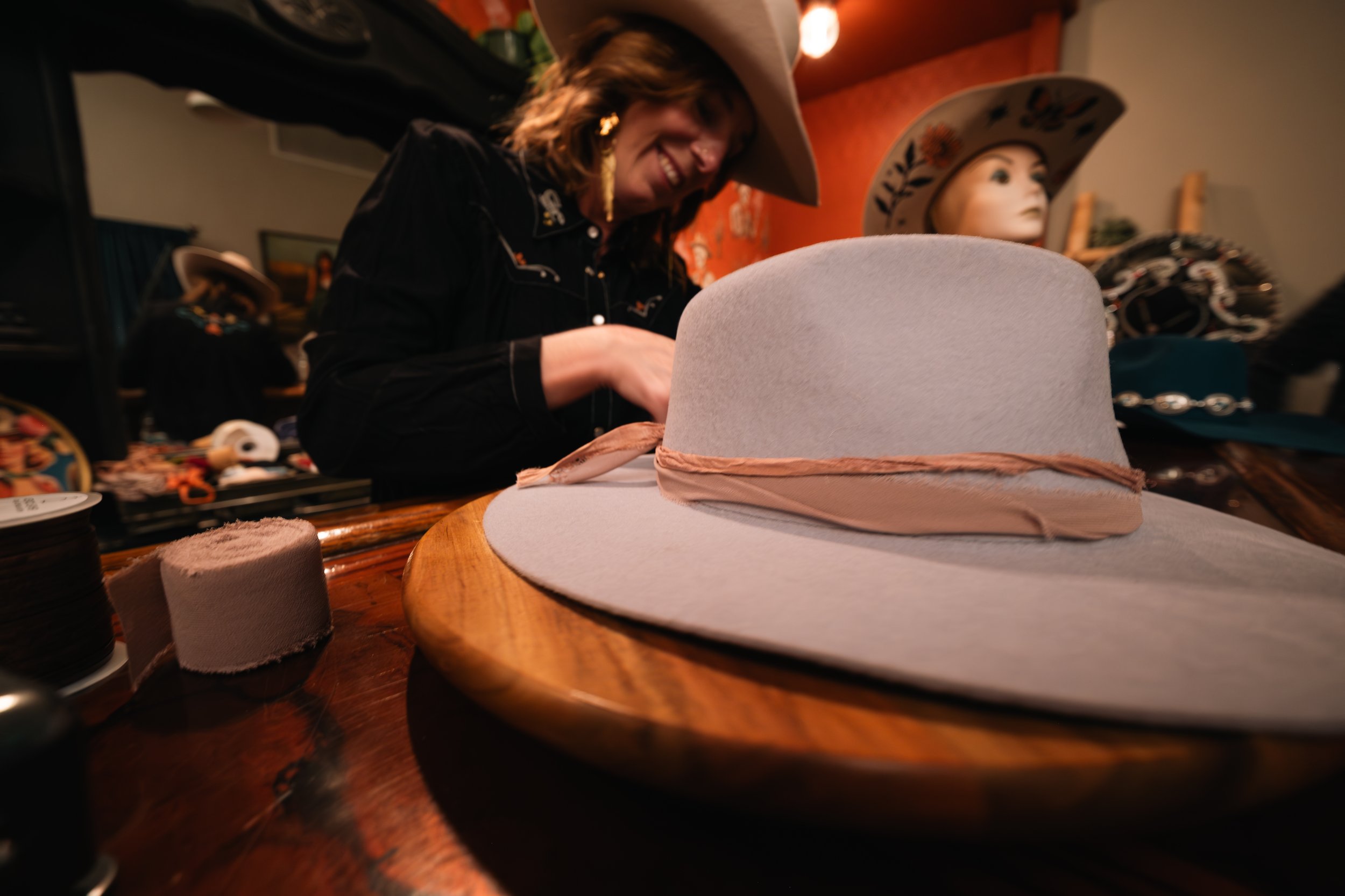 A woman smiling while trying on a white cowboy hat at a shop that sells hats and accessories, with display mannequins and various merchandise in the background.