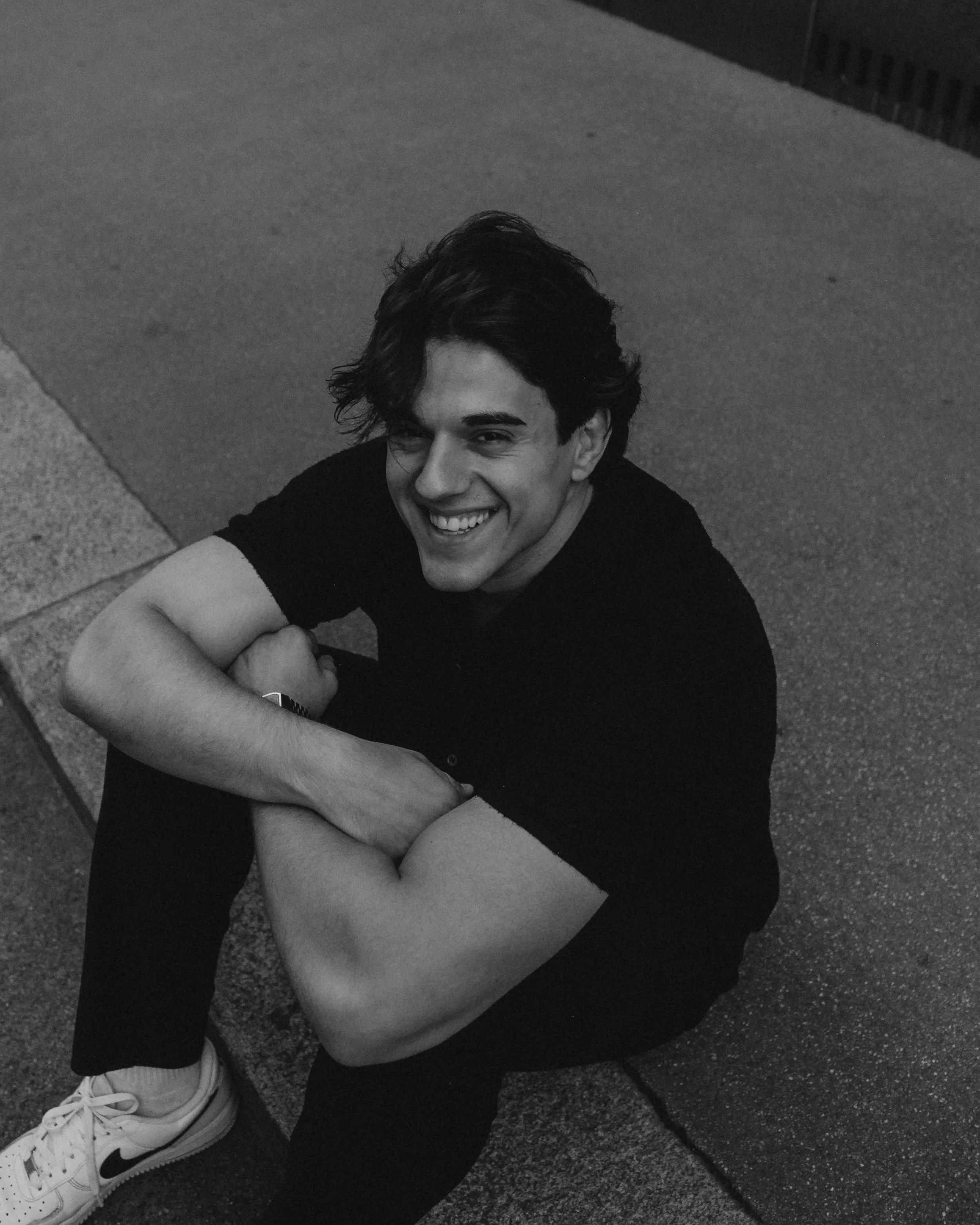 A young man with dark, curly hair sitting on the sidewalk, smiling and flexing his arm.