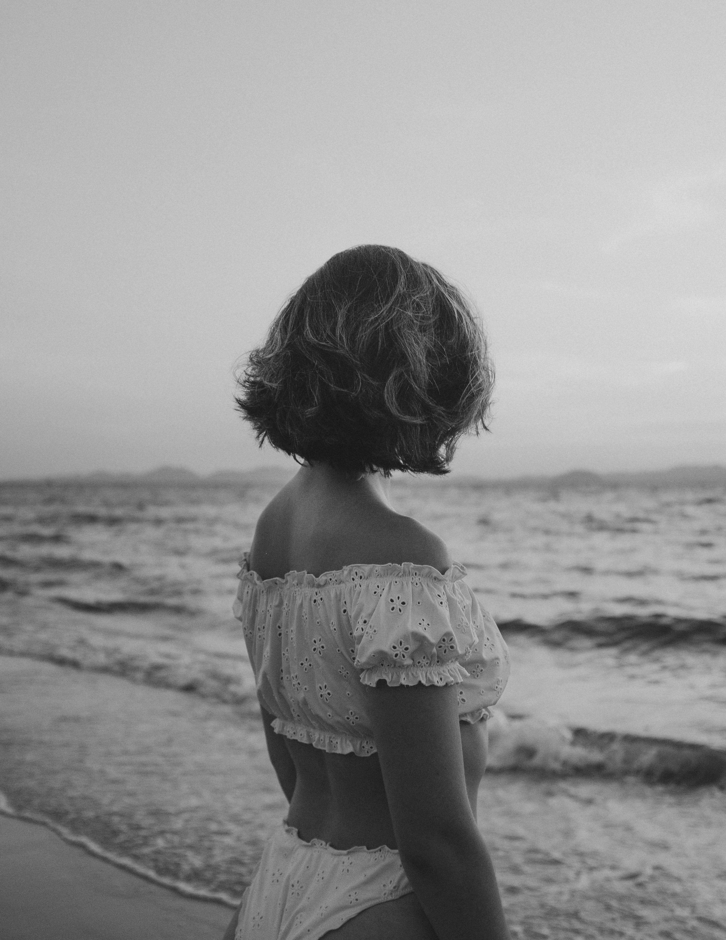 A woman with short wavy hair standing on a beach, looking at the water, wearing an off-the-shoulder top and matching bottoms.