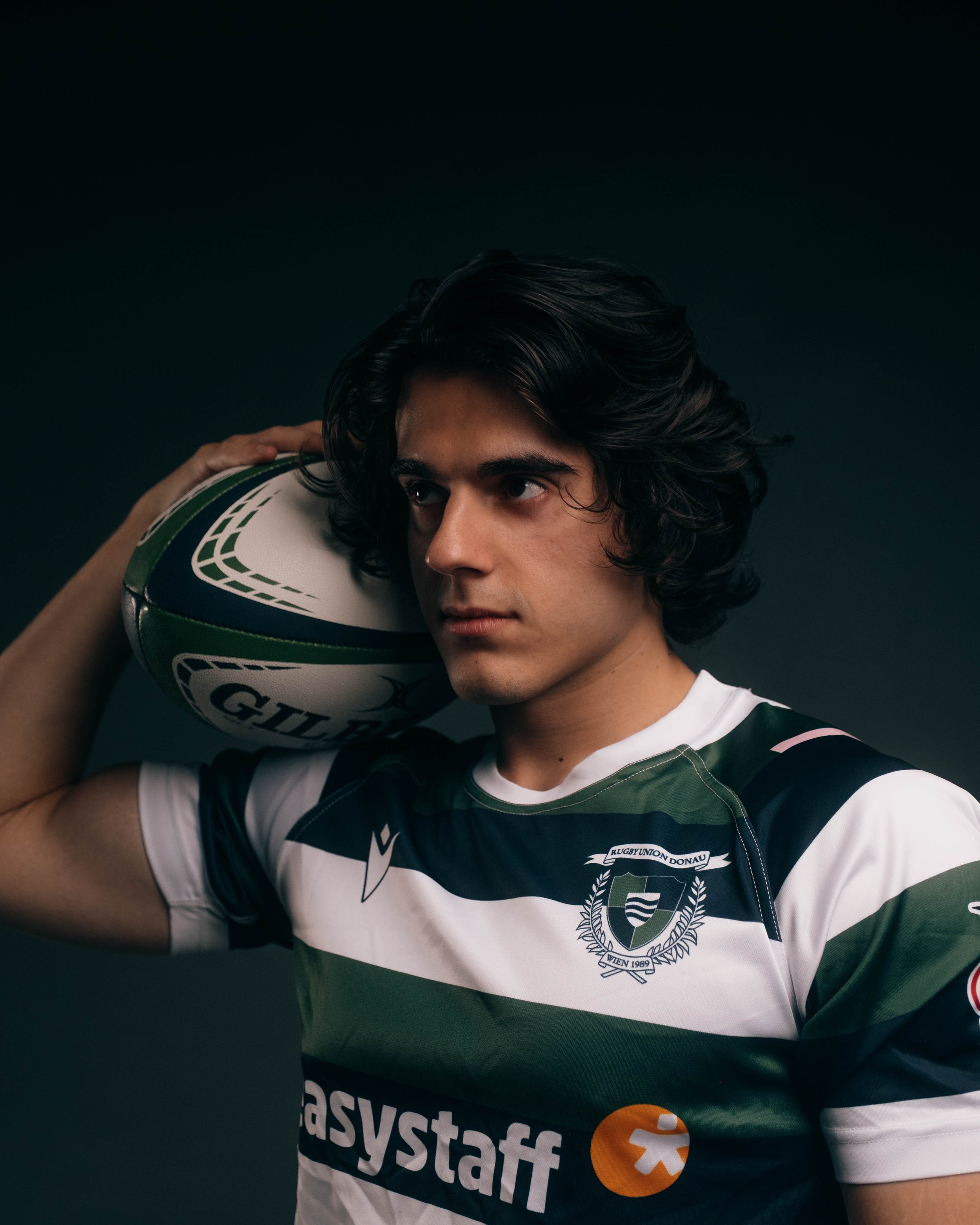 A young male rugby player with long black hair holding a rugby ball on his shoulder, wearing a dark green, white, and black striped jersey with a crest and the sponsor's logo, against a dark background.