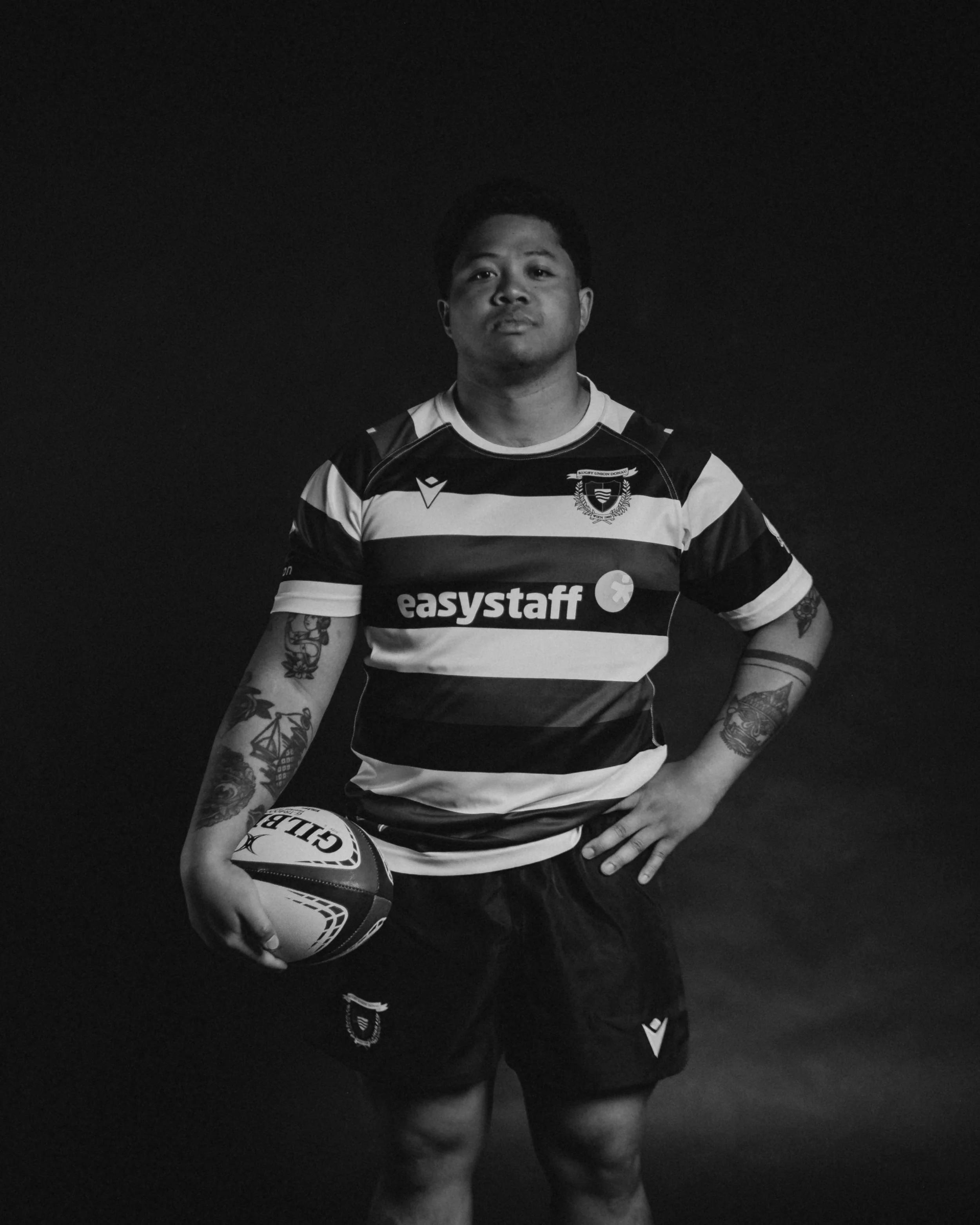 A male rugby player wearing a striped jersey with the words 'easystaff' on the front, holding a rugby ball, and standing against a dark background.
