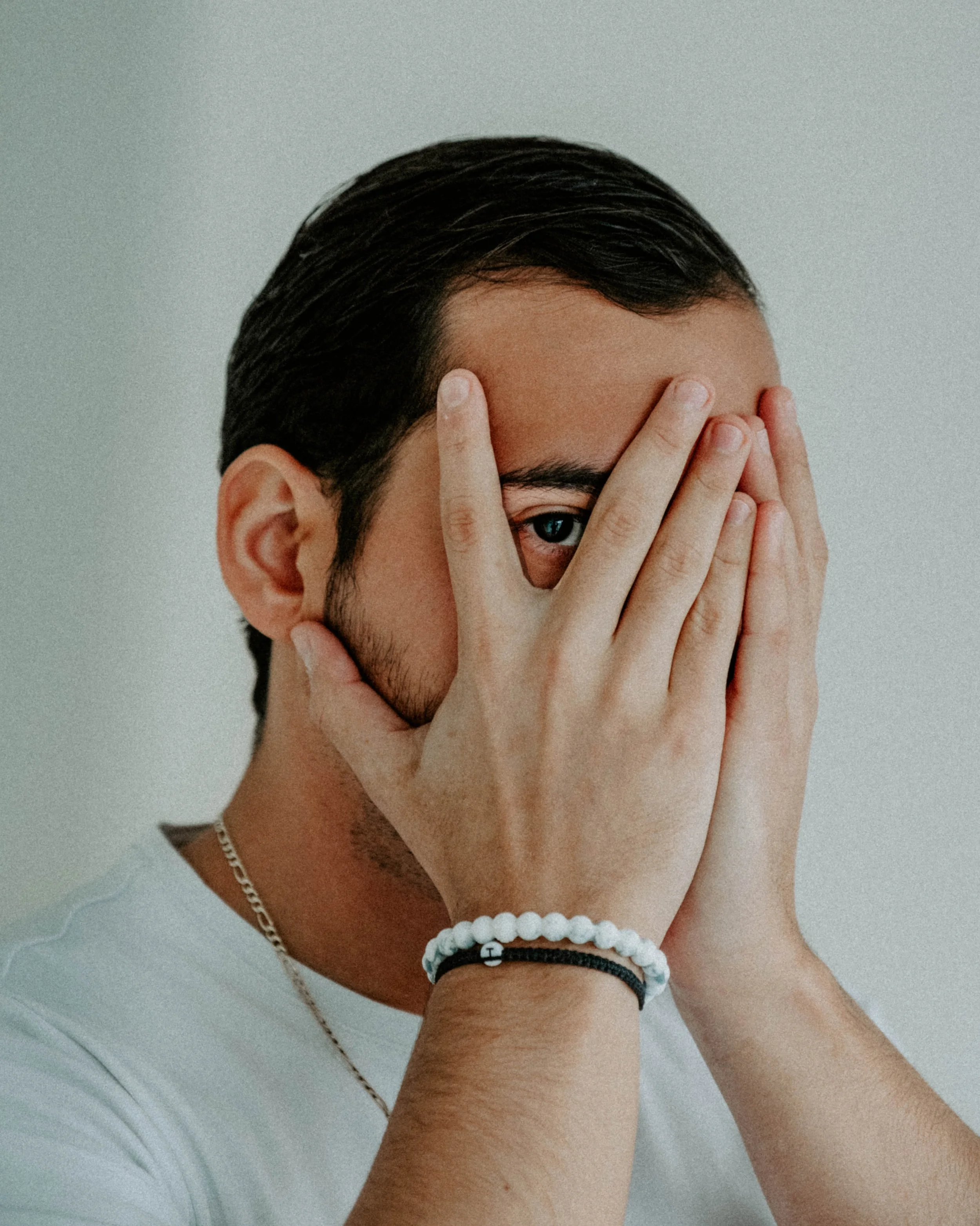 A young man with dark hair and light skin covering half his face with his hand, revealing one blue eye, against a plain background.