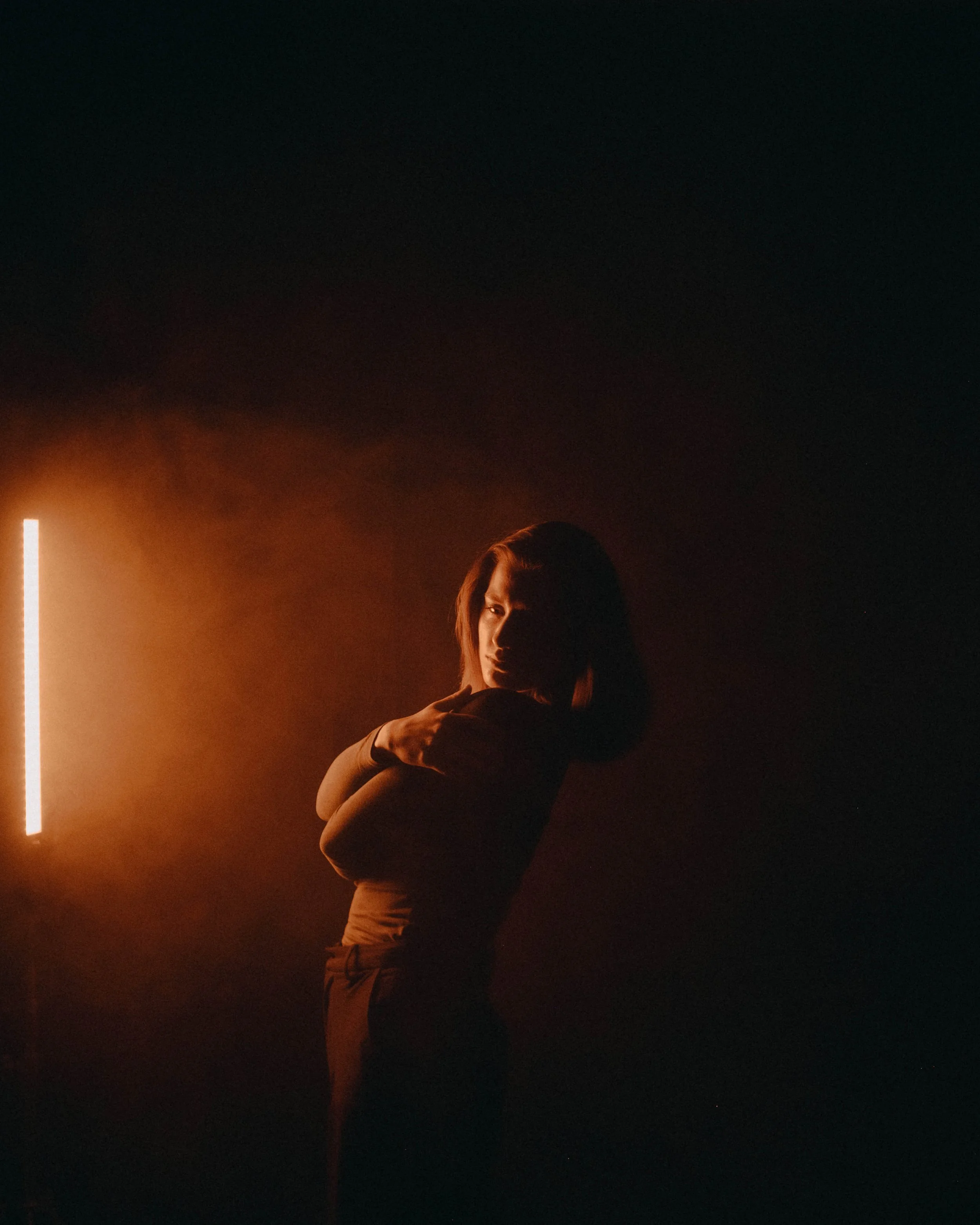 A woman with shoulder-length hair stands with arms crossed in a dark room lit by a single warm light on the left.