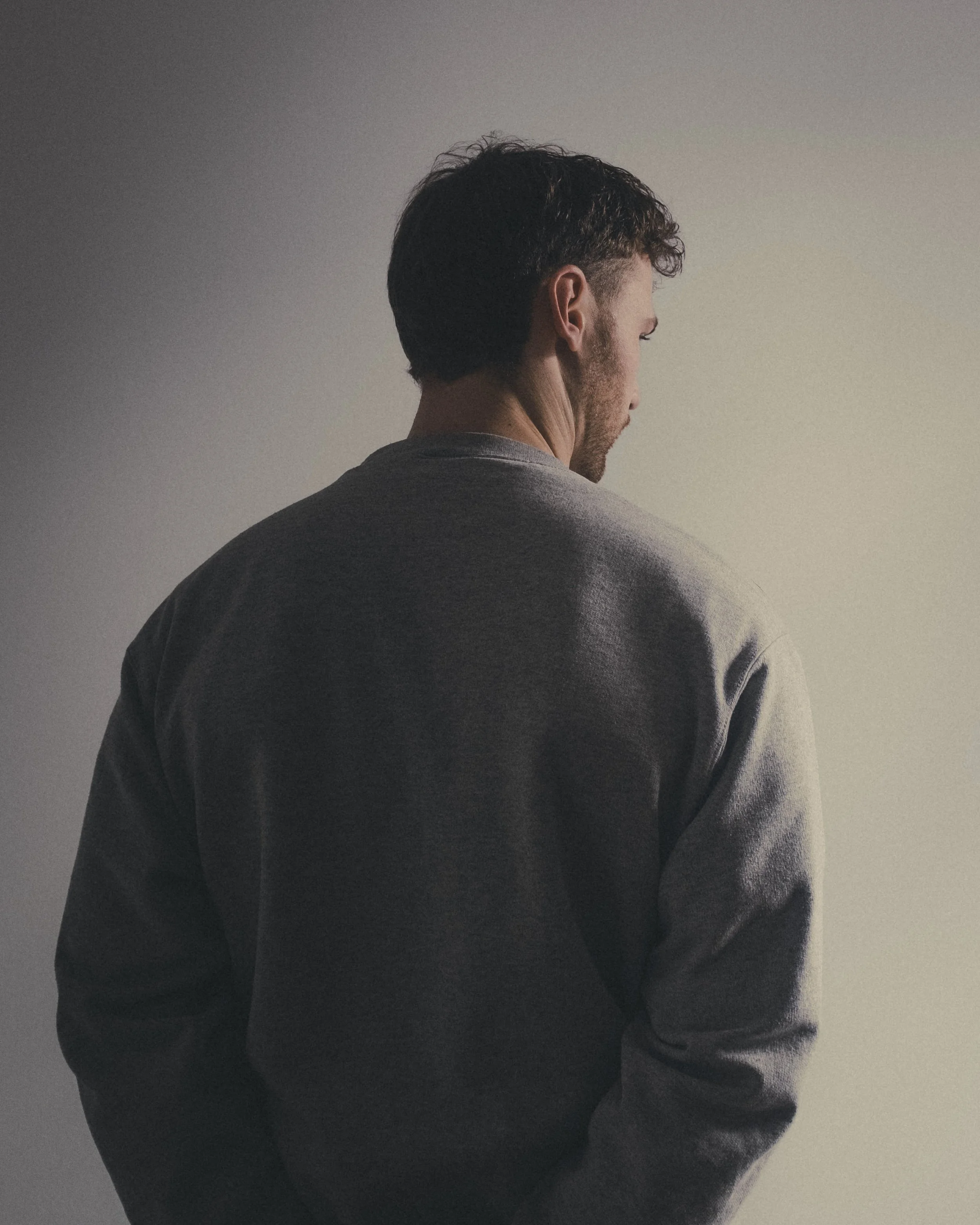 A man with dark hair and a beard, seen from behind, wearing a gray sweater, standing near a plain light-colored wall.