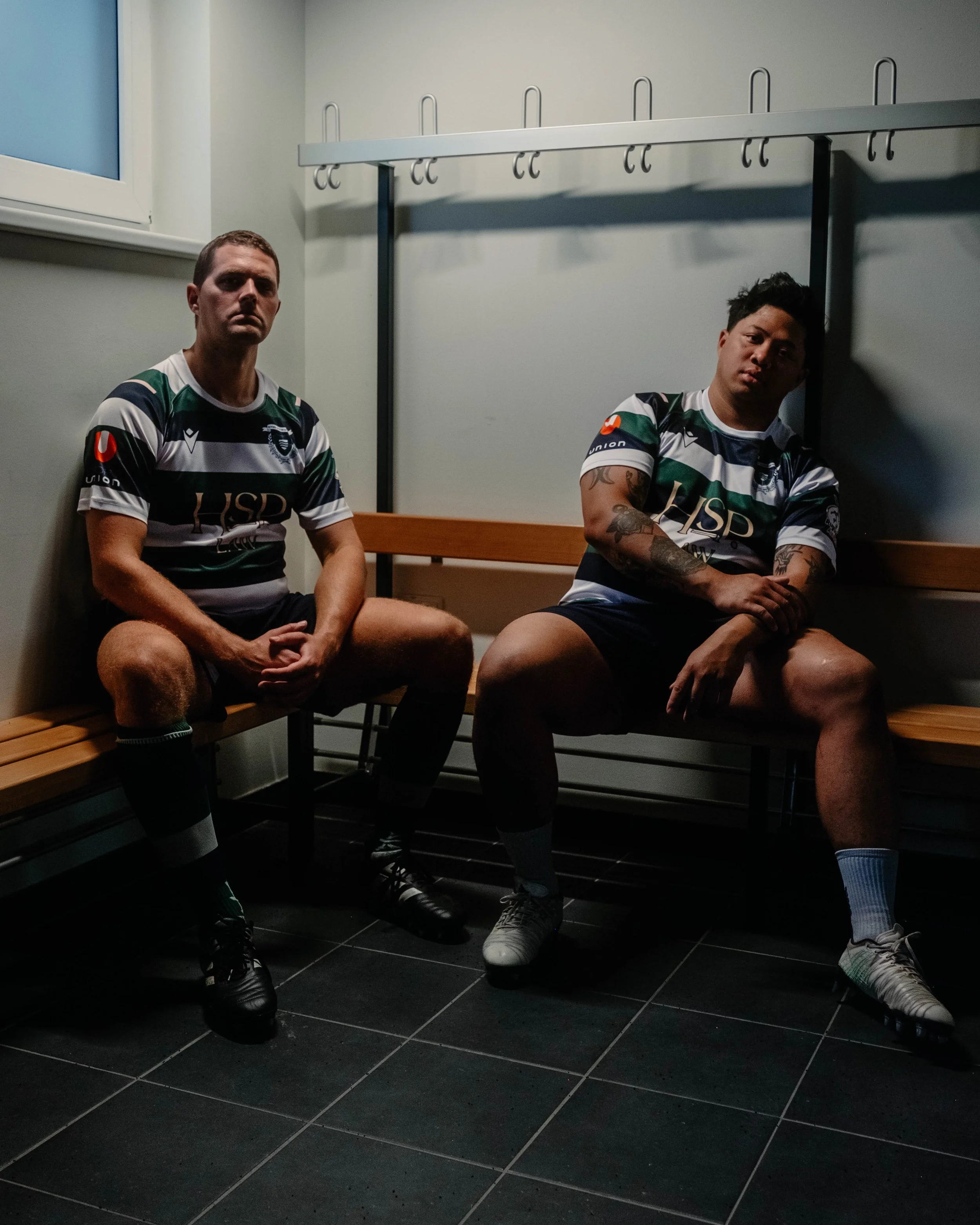 Two male soccer players sitting on a wooden bench in a locker room, wearing black and white striped jerseys, black shorts, and soccer cleats.