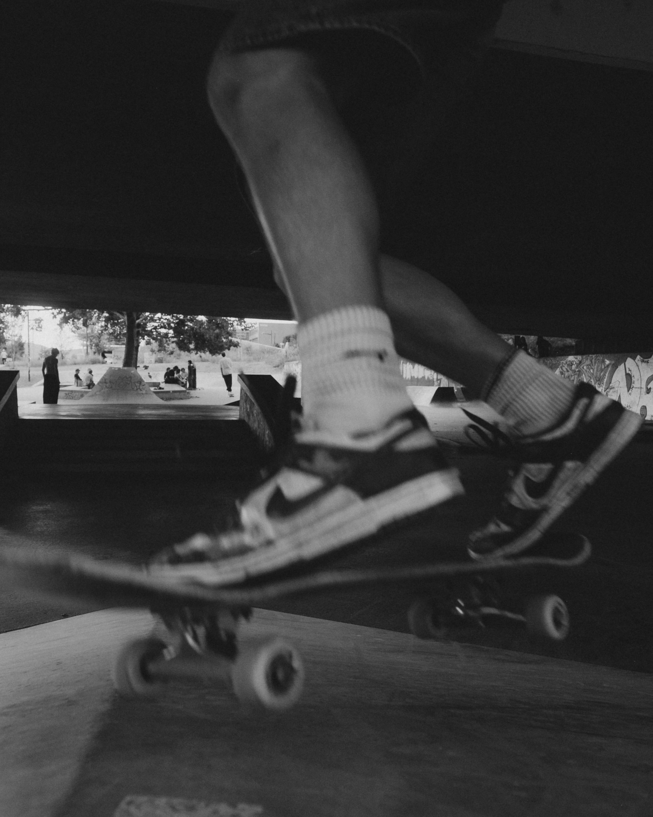 A person skateboarding at a skate park, with their legs and skateboard visible as they perform a trick underneath a structure, with people and trees in the background.