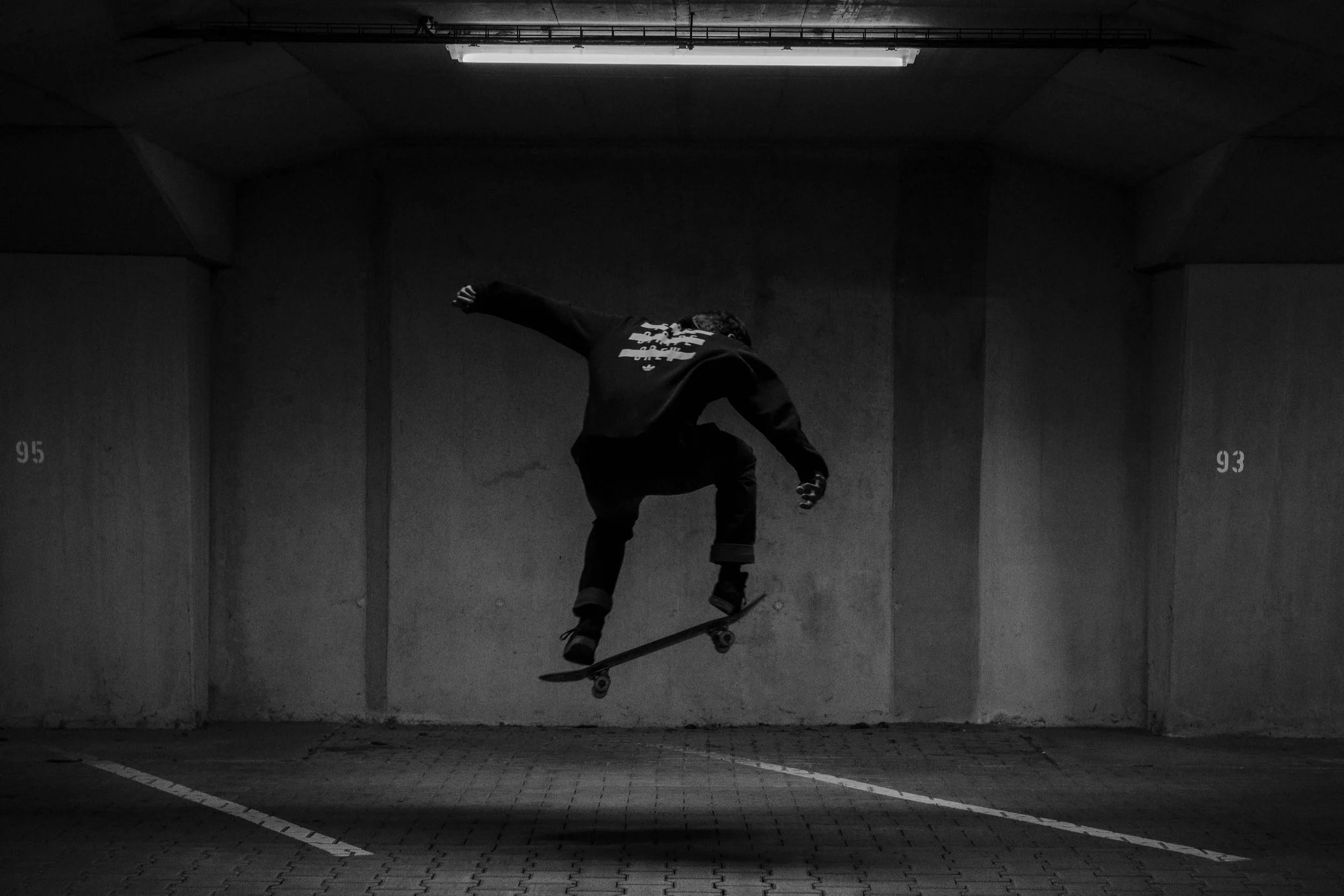Skateboarder performing a trick in a parking garage, captured in black and white, with numbered parking spaces 95 and 93 visible on the walls.