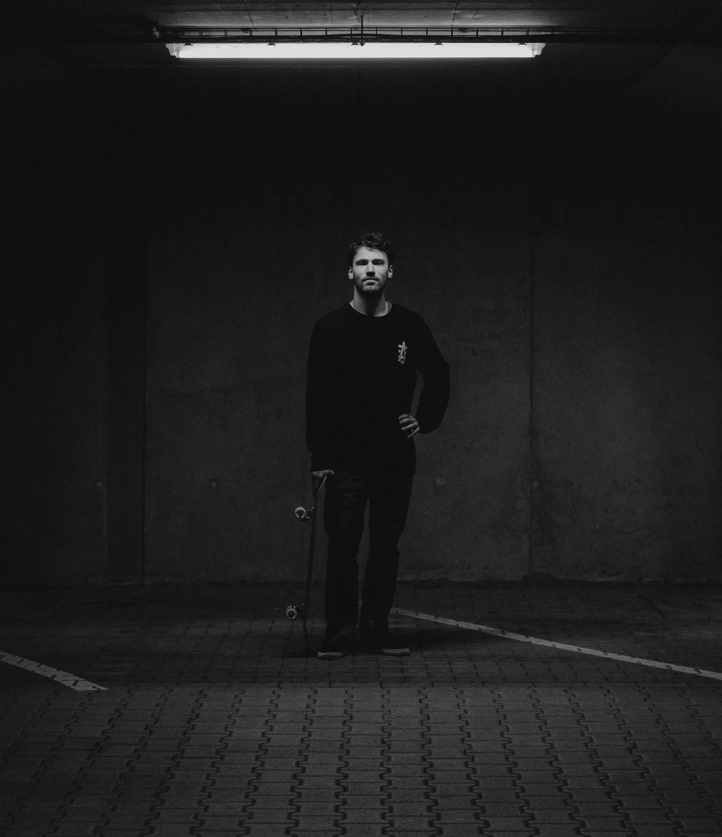 A young man stands in an underground parking garage, holding a skateboard in one hand, with a confident pose, wearing a black long-sleeve shirt and black pants, illuminated by a single overhead fluorescent light.