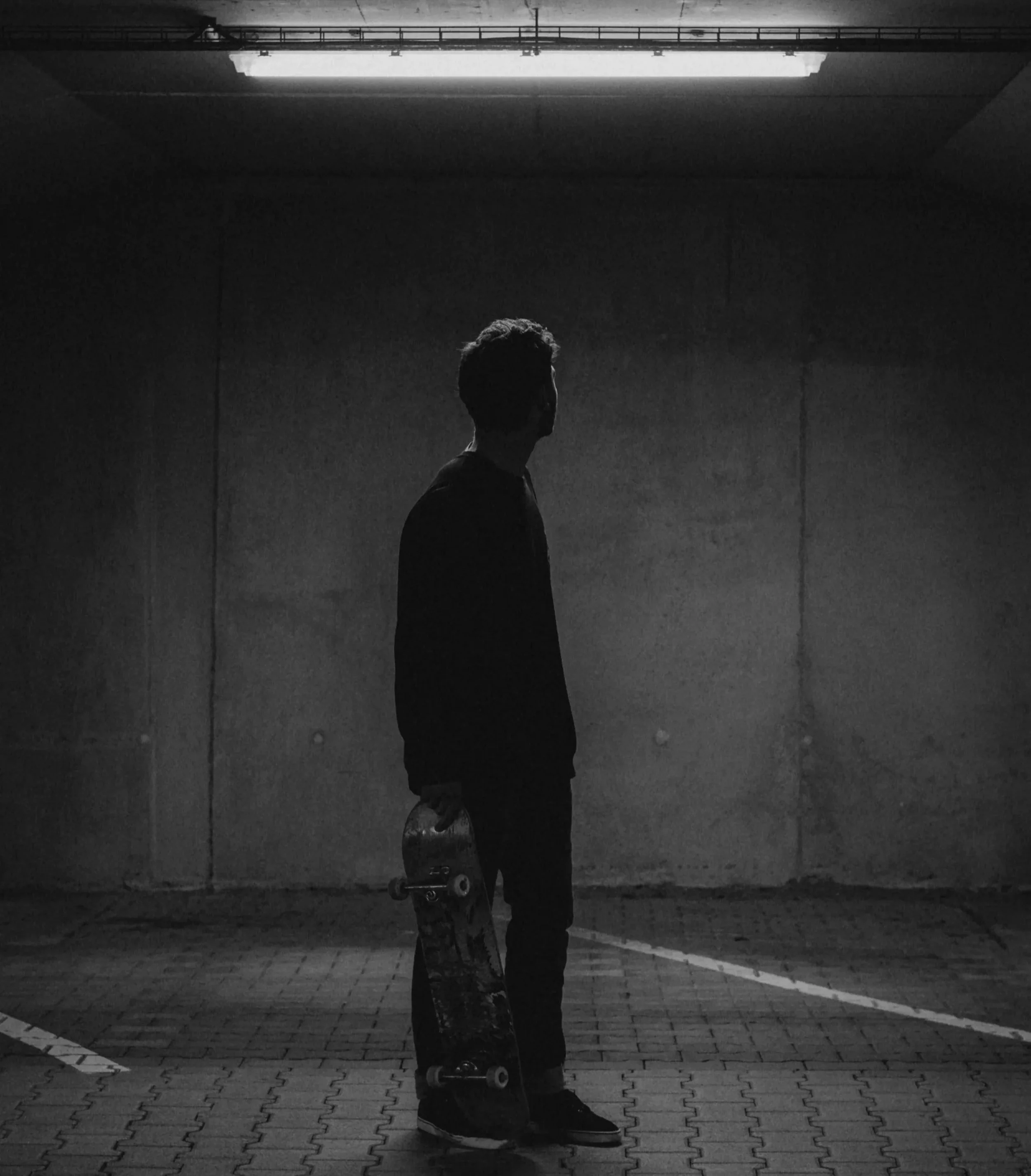 A person standing in a dimly lit outdoor parking garage holding a skateboard, silhouetted against an illuminated ceiling.