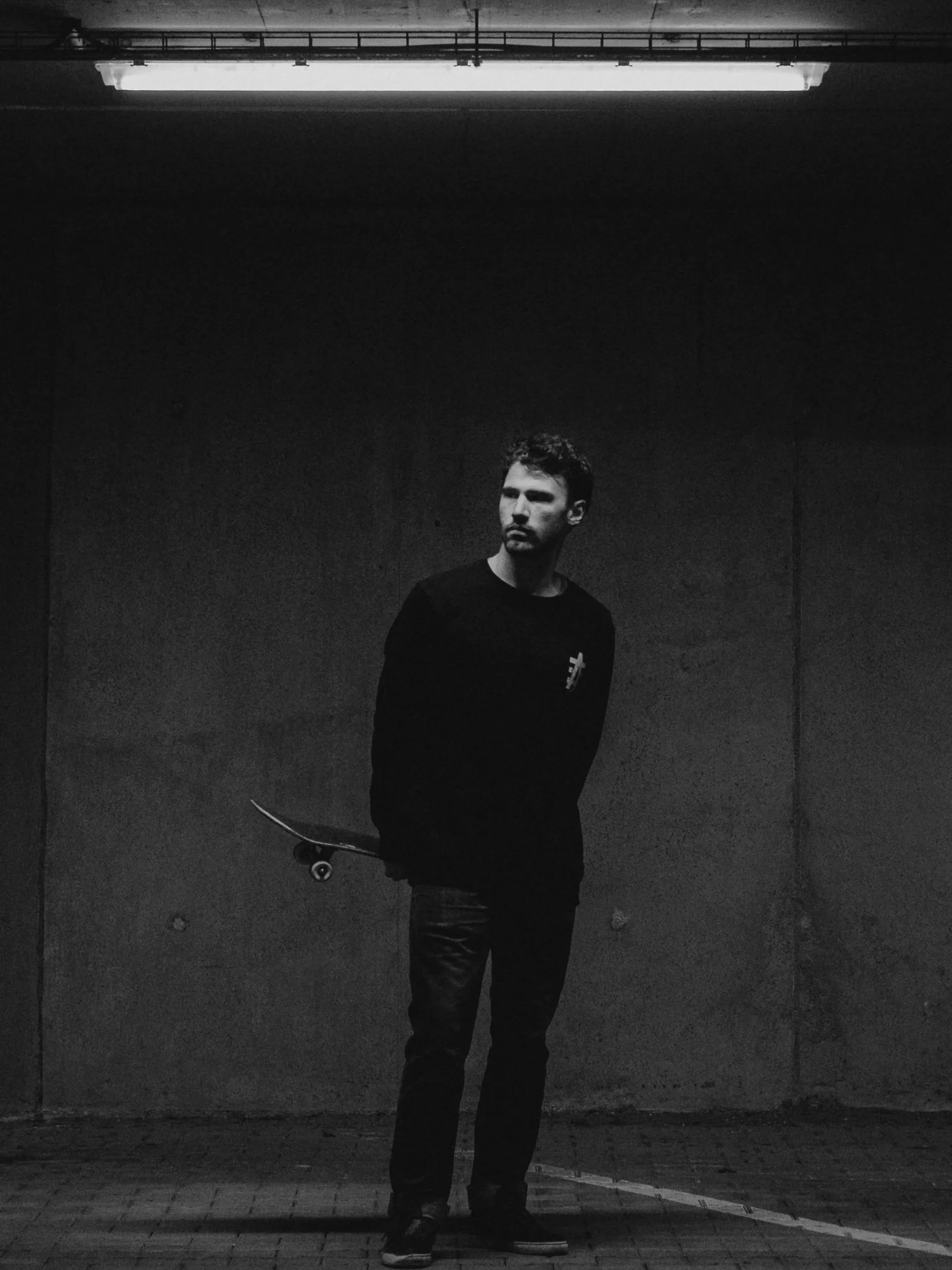 A man standing in front of a plain wall, holding a skateboard behind his back, in a dimly lit space with a fluorescent light overhead.