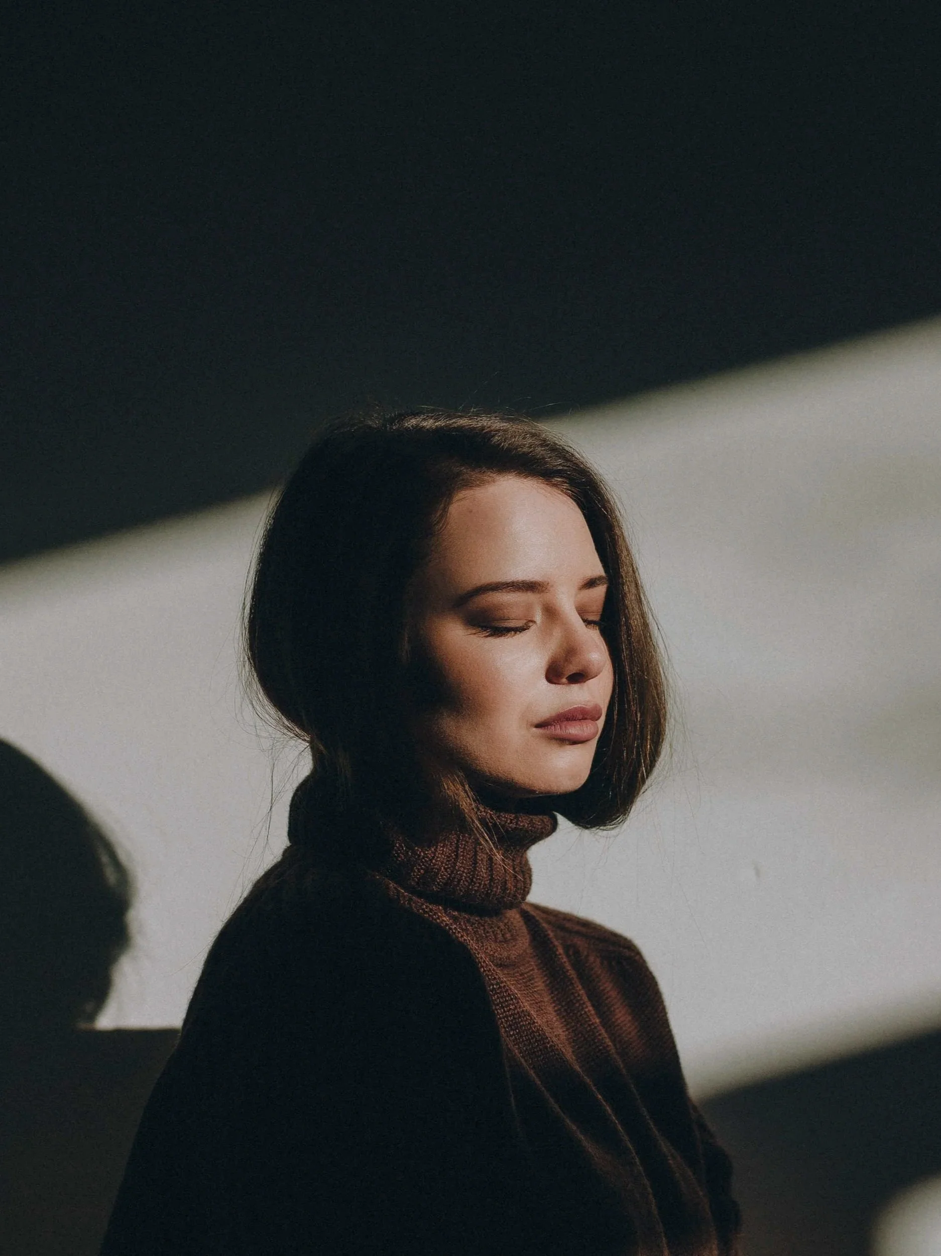 A woman with shoulder-length brown hair wears a brown turtleneck sweater with her eyes closed, standing in front of a white wall with shadows cast across her face and background.