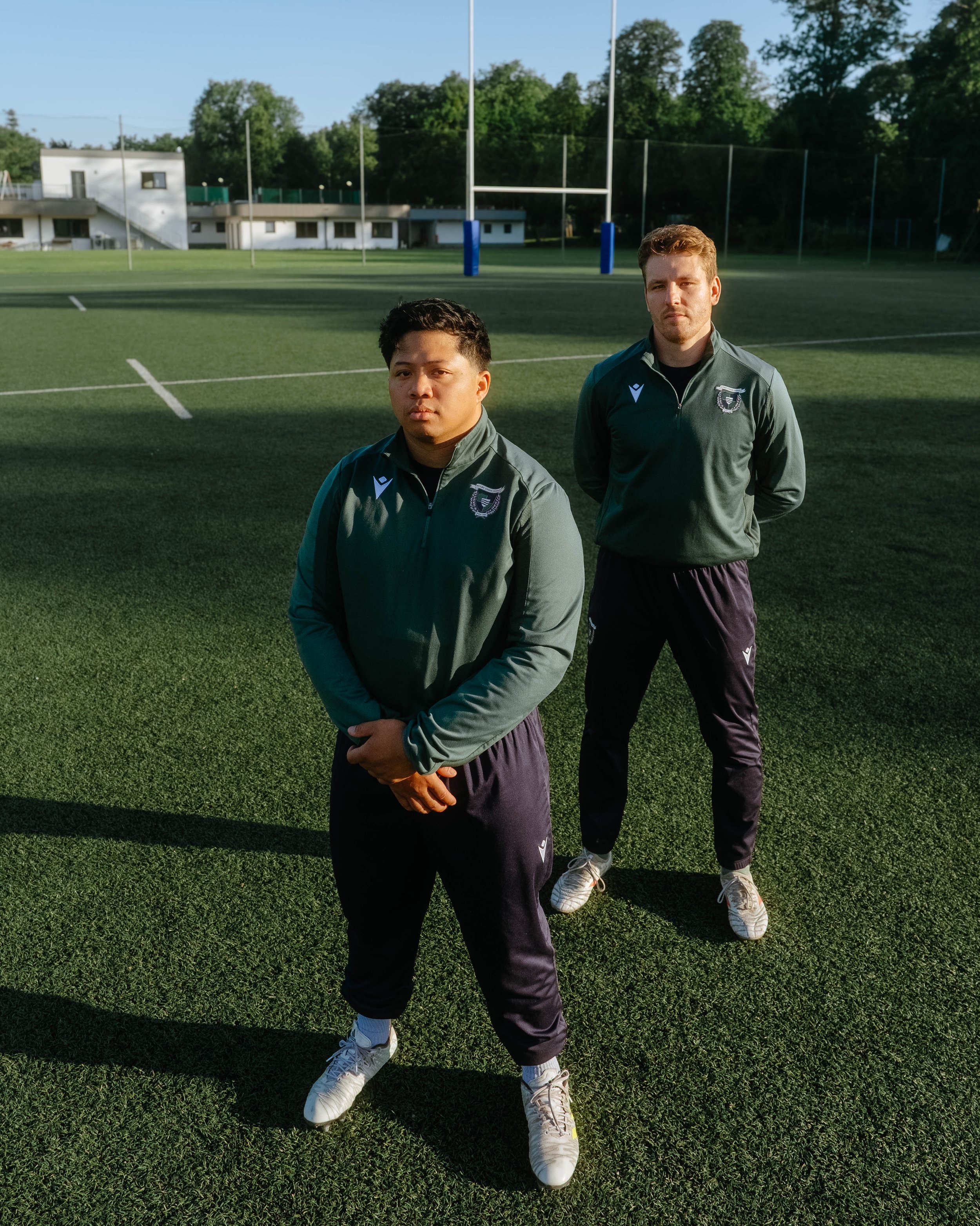 Two male soccer players standing on a green field, wearing matching green sports jackets and black athletic pants, with goalposts and trees in the background.