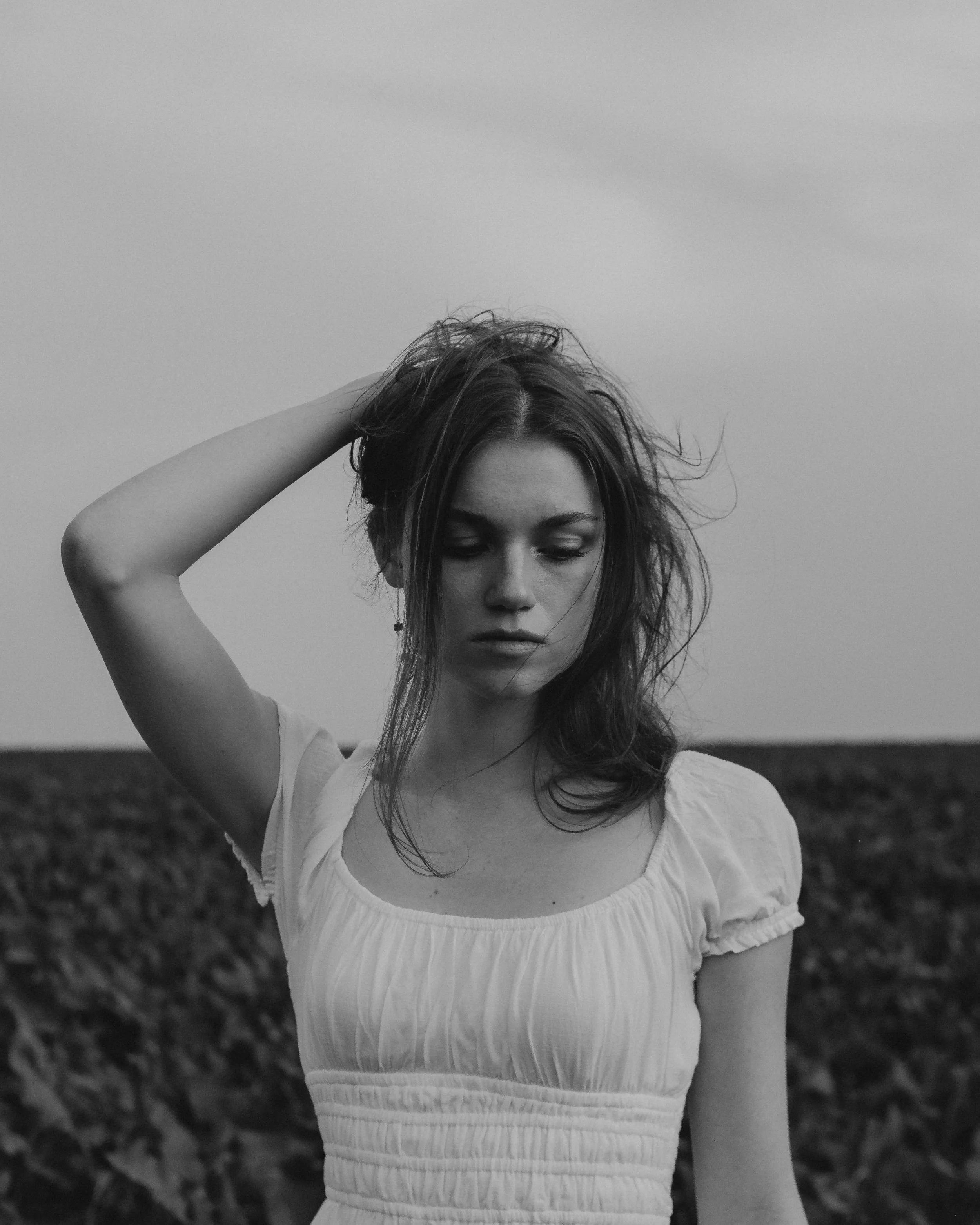 A black and white photo of a young woman with long hair, wearing a light-colored dress, standing outdoors with a cloudy sky in the background.