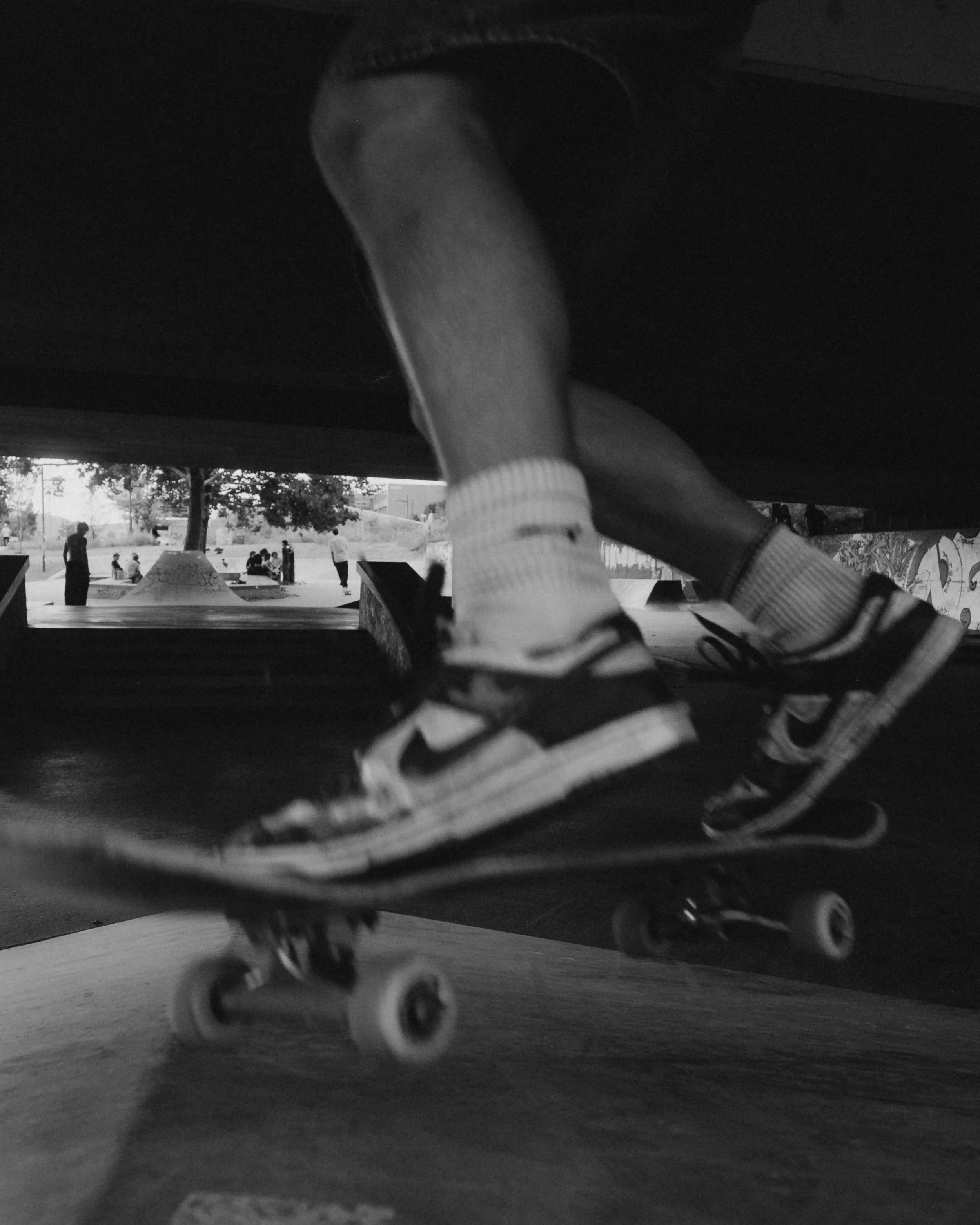 Child's legs and feet on a skateboard, viewed from below in a black-and-white photo at a skate park with children and skateboarders in the background.