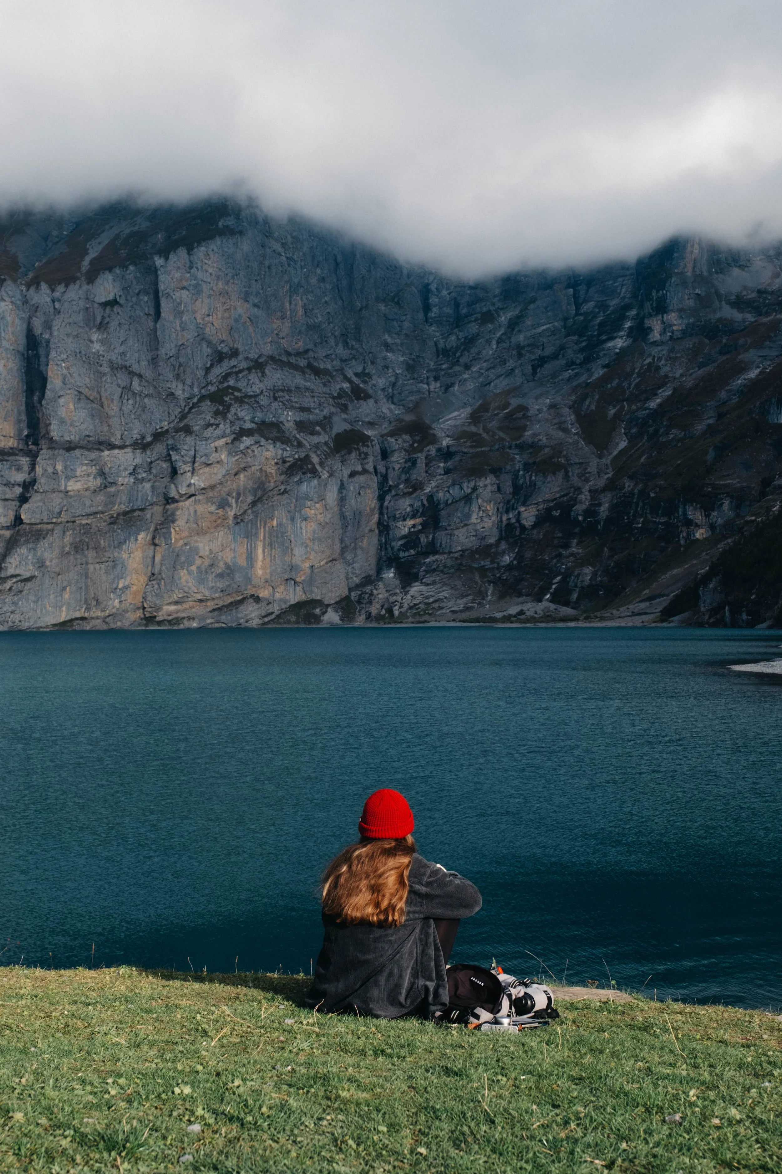 A woman with long hair, wearing a red beanie and dark jacket, sitting on the grass by a lake with mountains in the background, cloudy sky overhead.