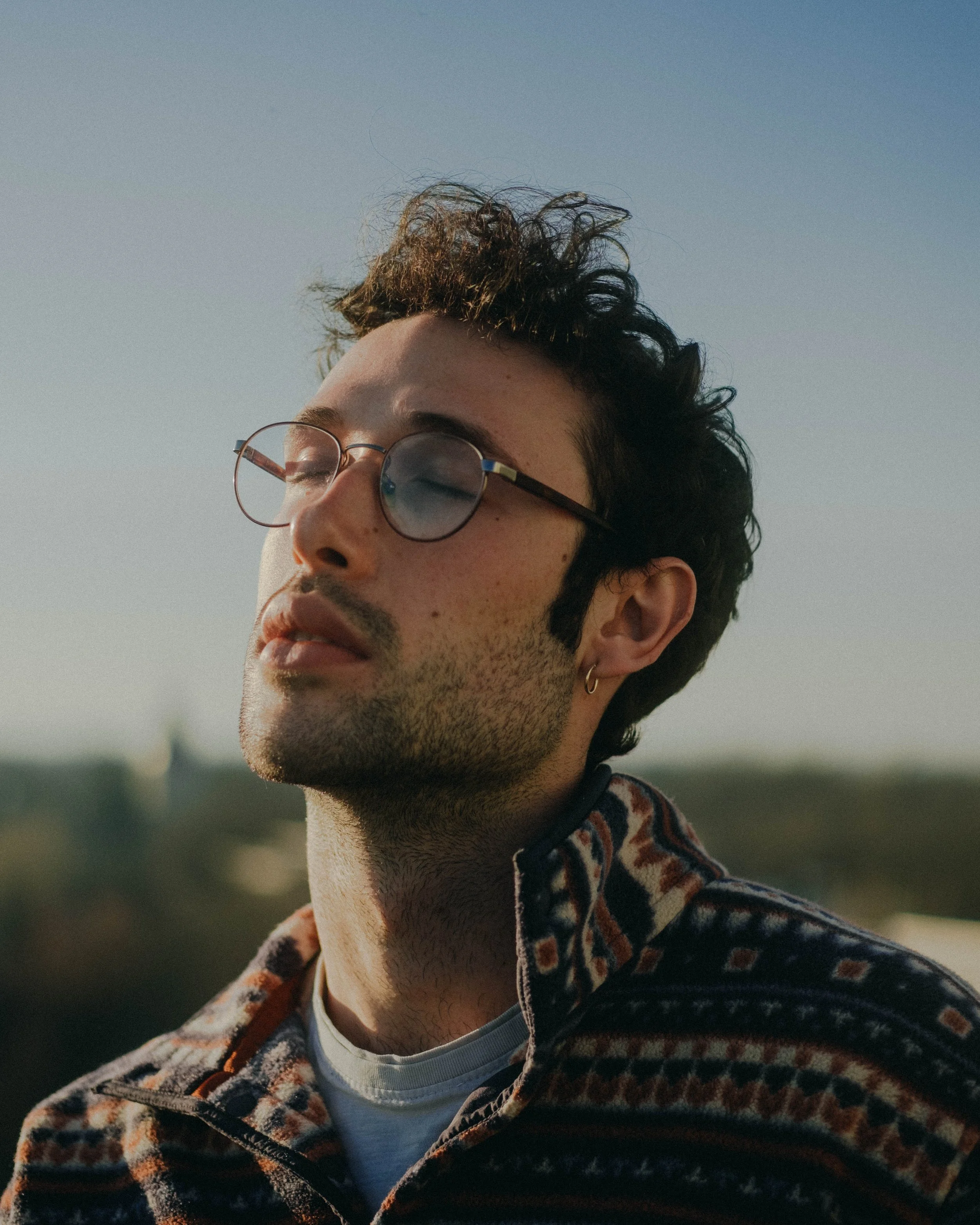 Close-up of a young man with curly dark hair, wearing round glasses, earrings, a patterned zip-up jacket, and a white shirt underneath, with a blurred outdoor background and his eyes closed.