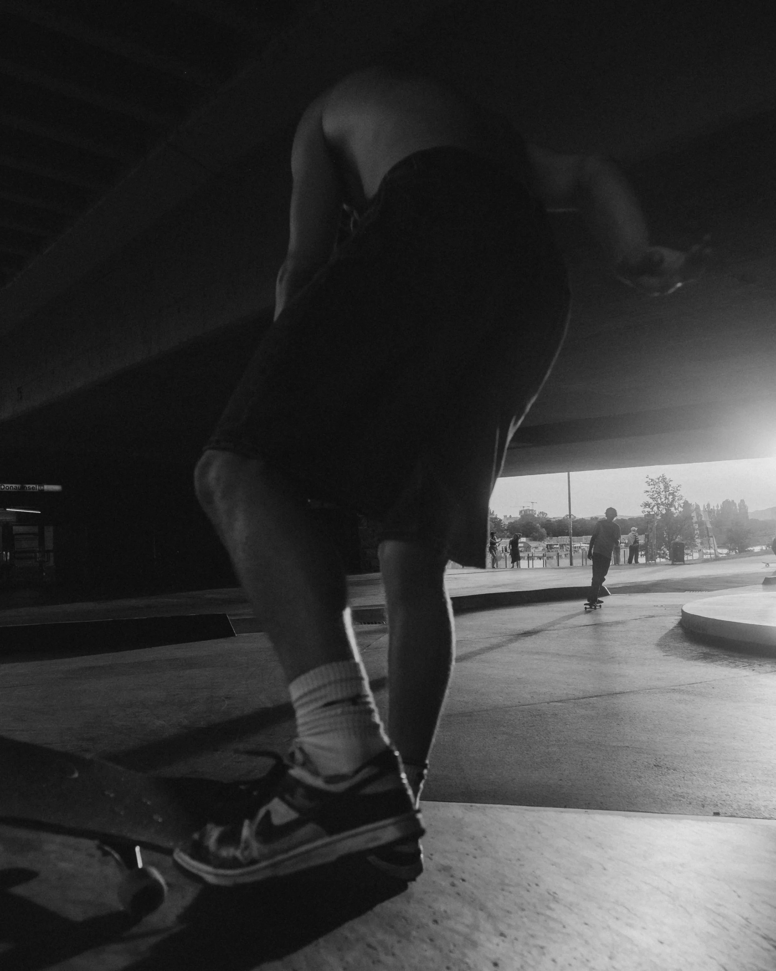 A person bending down on a skateboard under a bridge at sunset, with other people in the background.