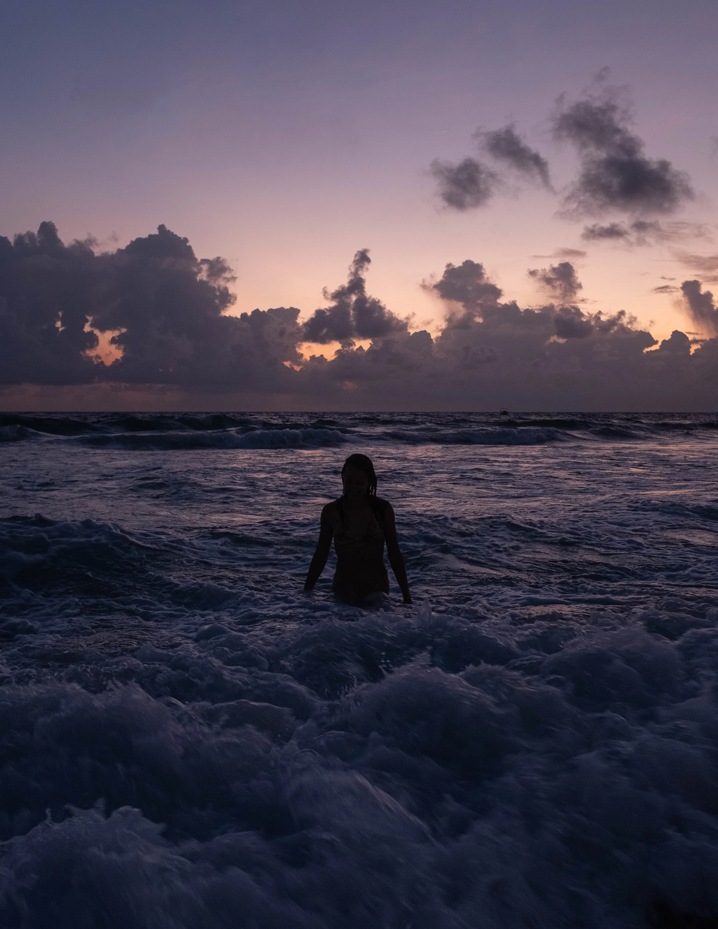 Silhouette of a person standing in the ocean during sunset with a colorful sky and clouds in the background.