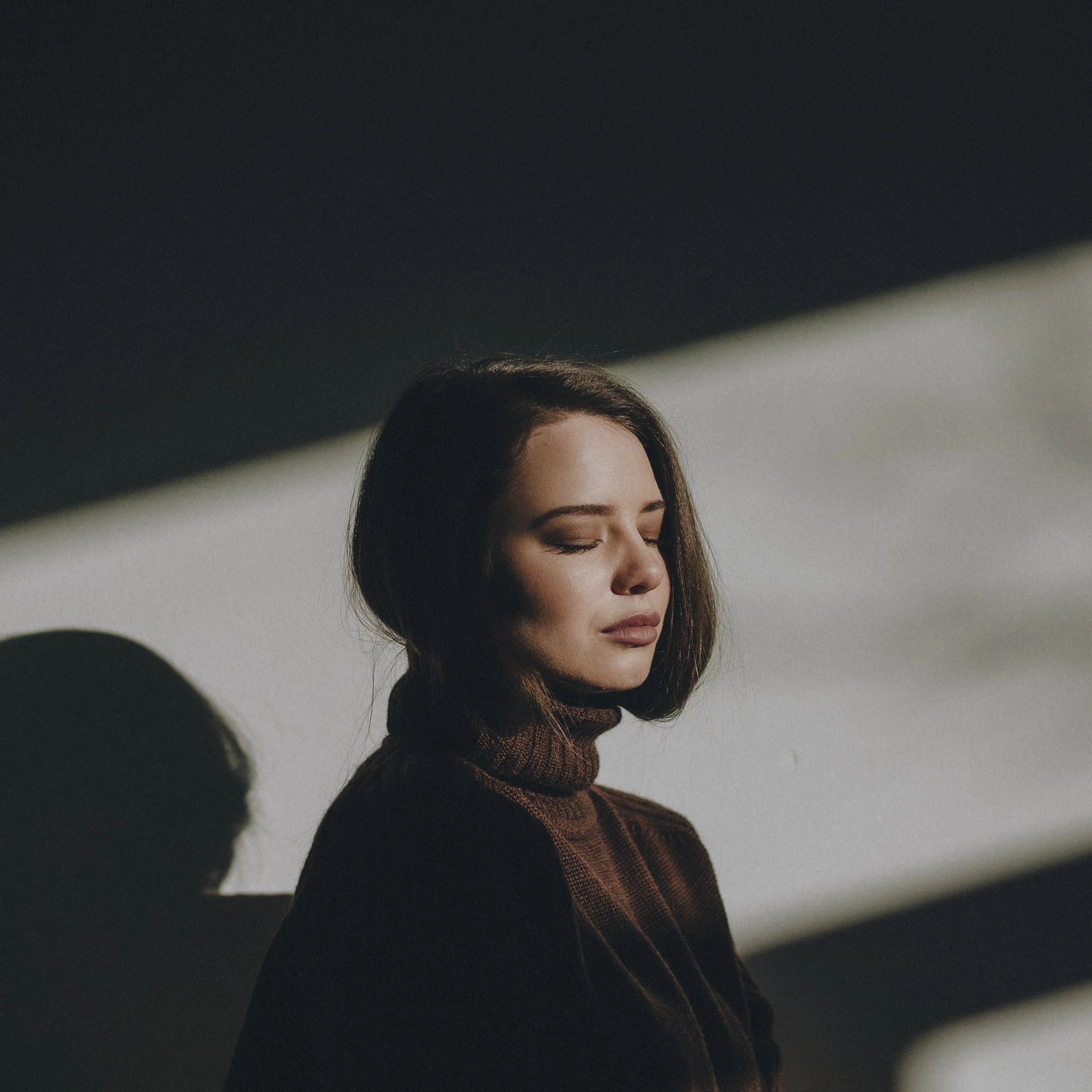A woman with shoulder-length brown hair wears a brown turtleneck sweater with her eyes closed, standing in front of a white wall with shadows cast across her face and background.