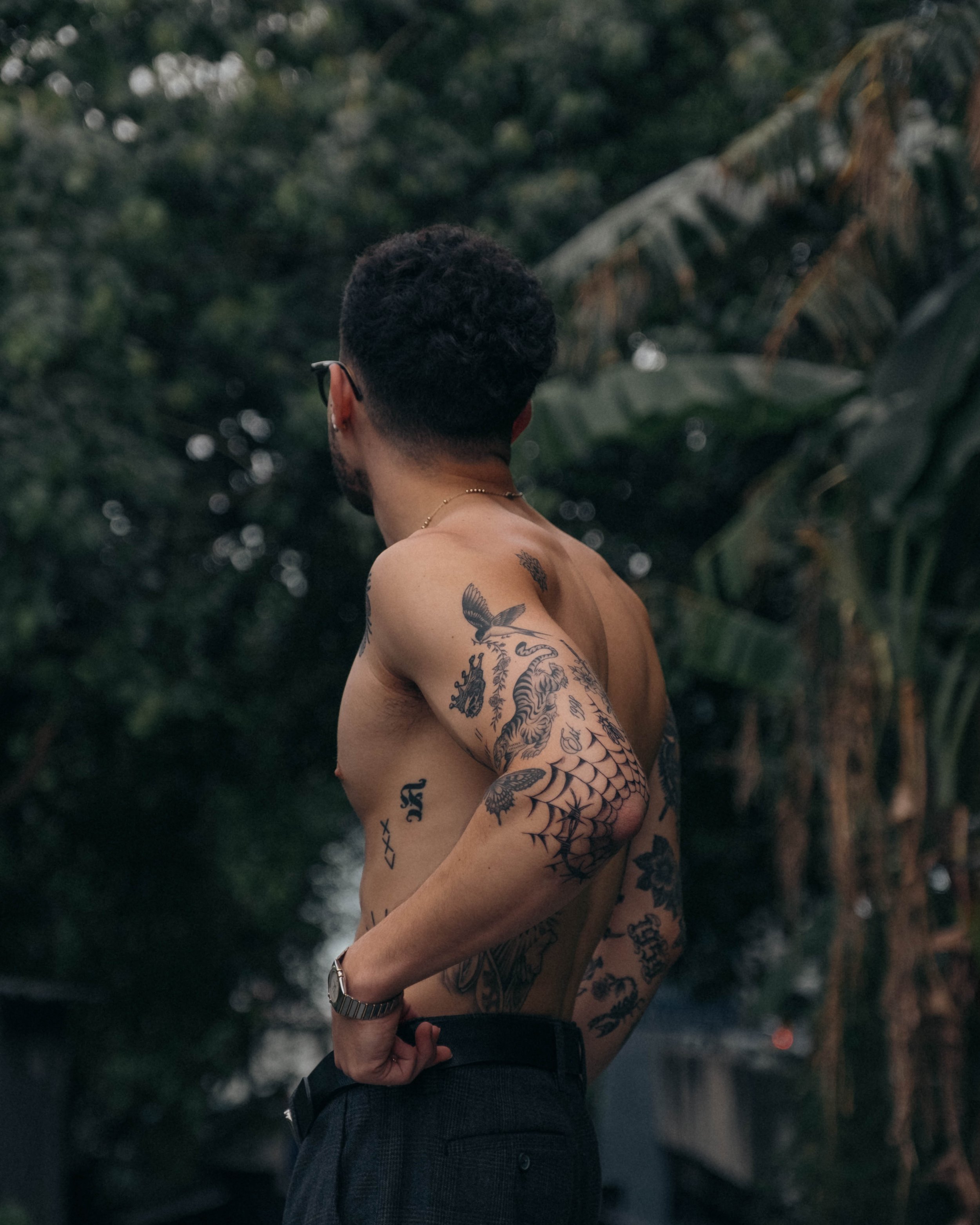 A shirtless man with tattoos on his back and arms, wearing glasses, standing outdoors with trees and foliage in the background.