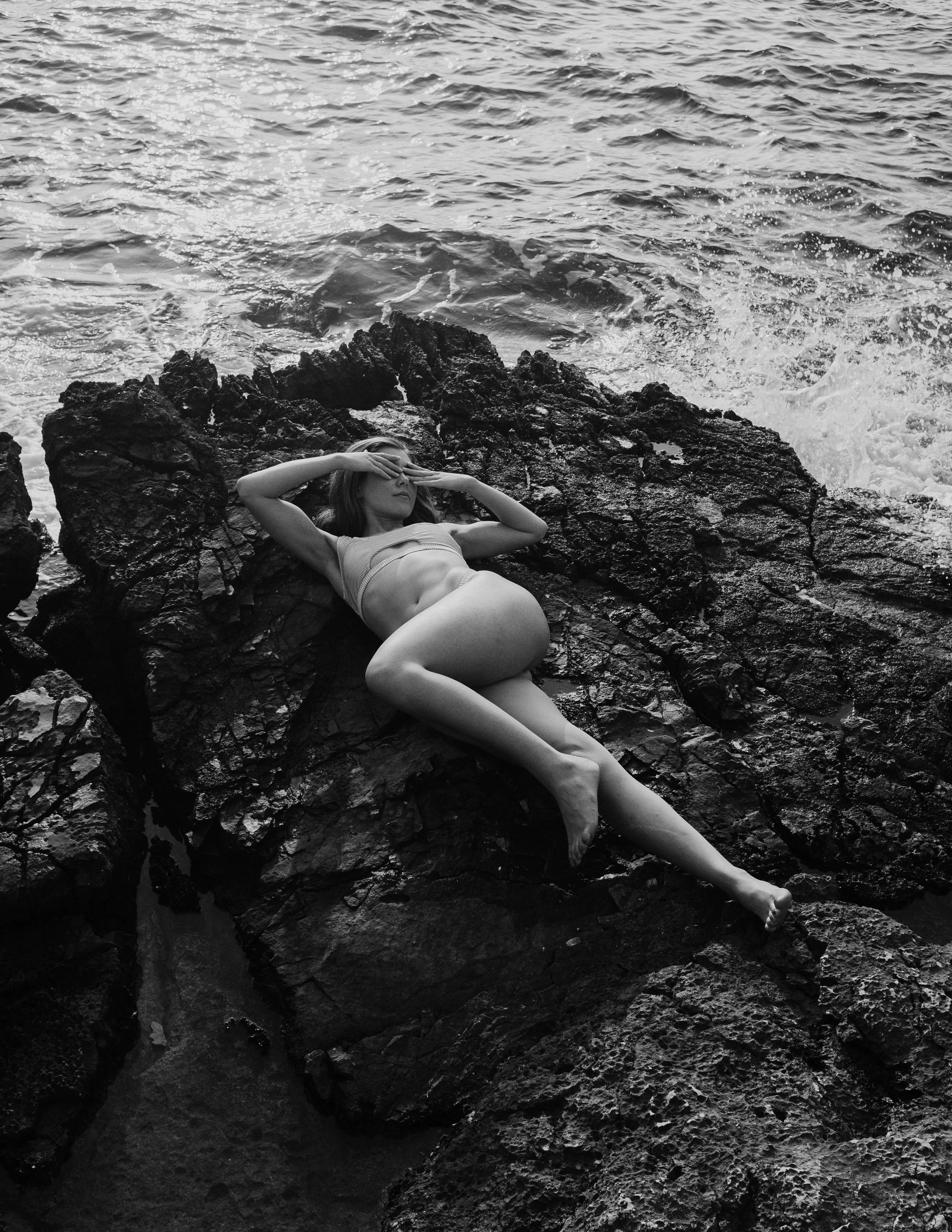 A woman in a swimsuit relaxing on rocks by the sea, shielding her eyes from the sun.