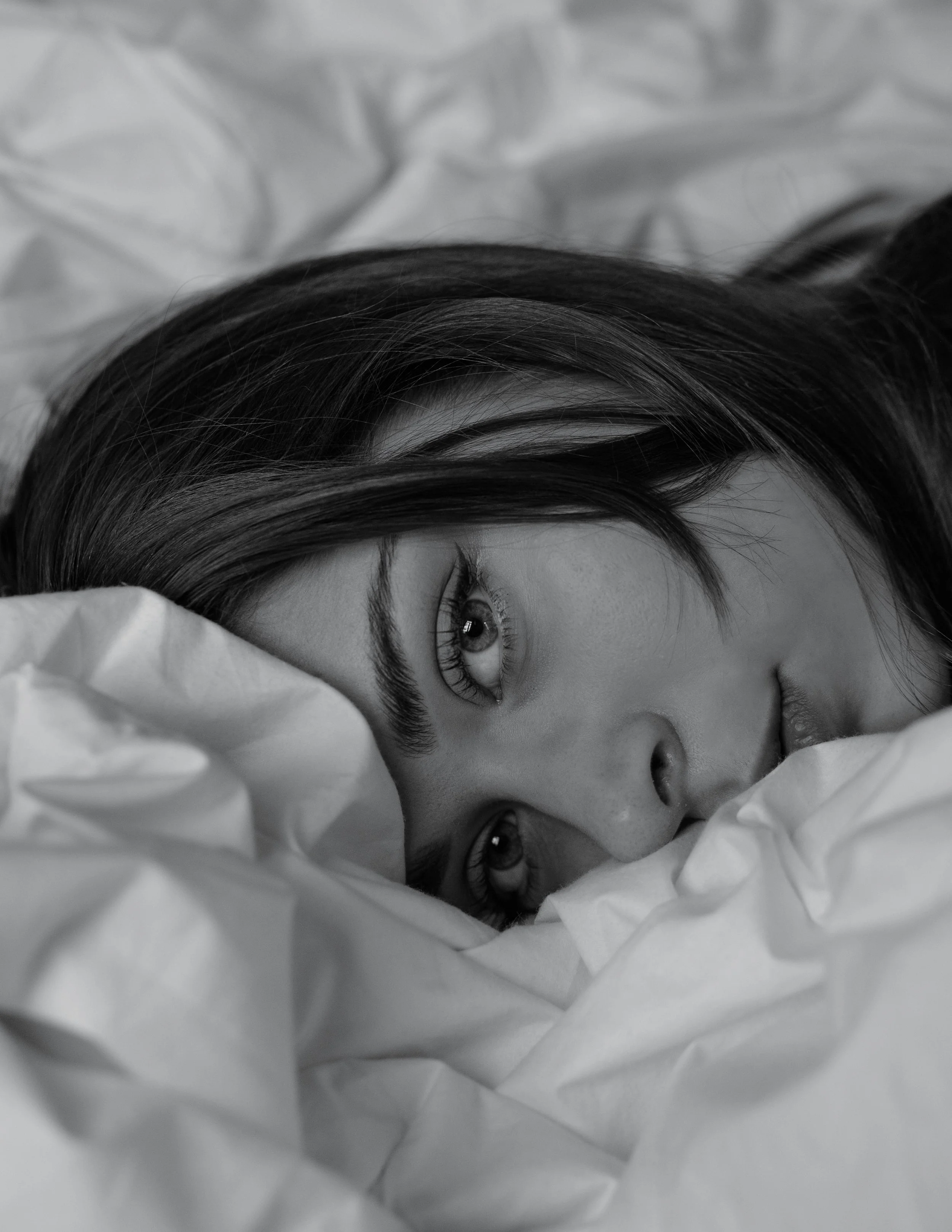 Close-up of a woman lying on crumpled bedsheets, with her face partially covered by the sheets, looking into the camera.