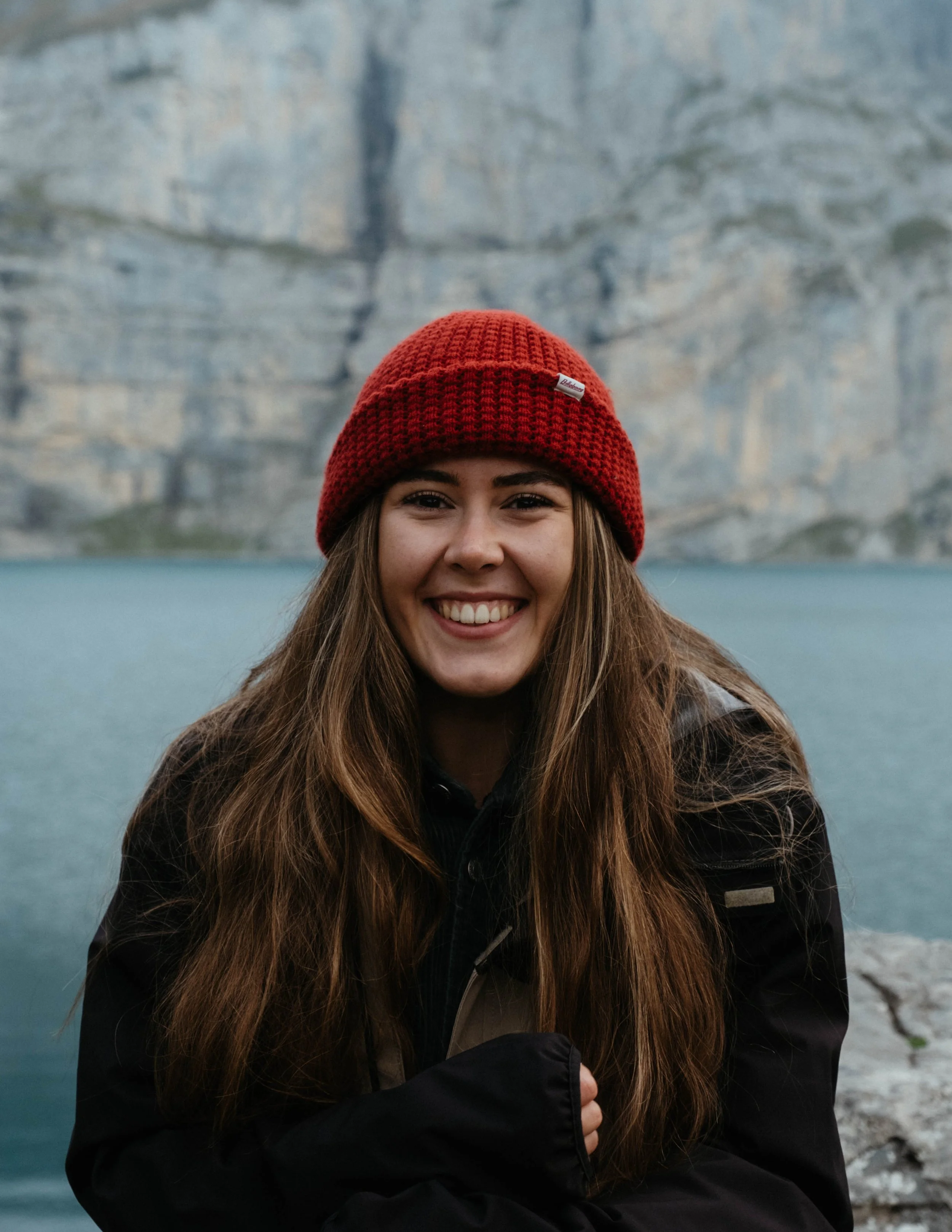 A young woman smiling outdoors near a lake with rocky cliffs in the background, wearing a red knit beanie and a black jacket.