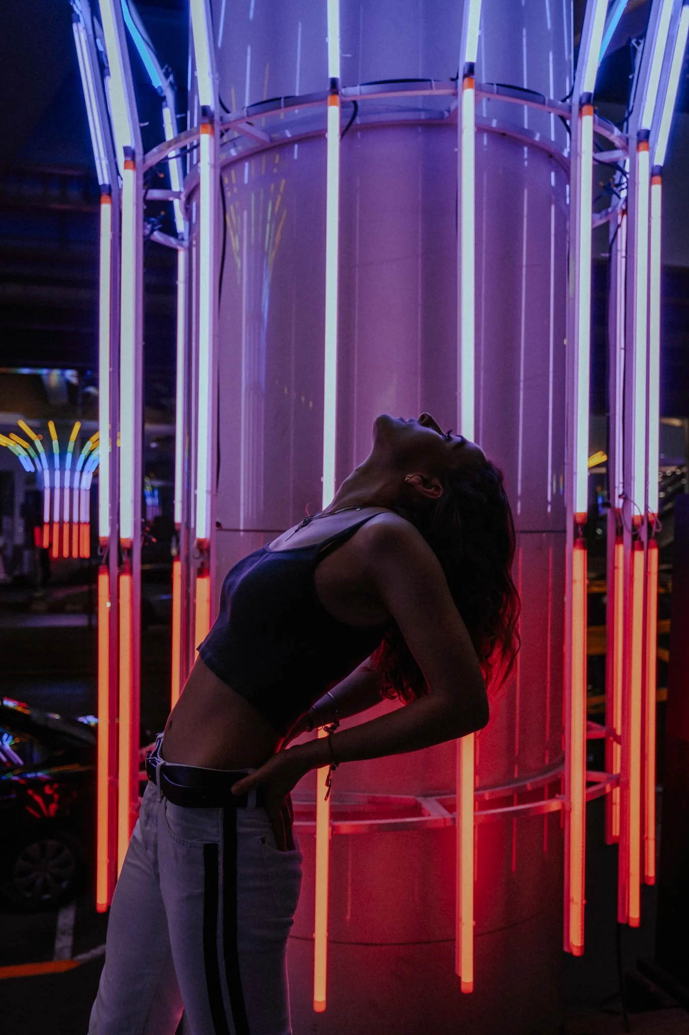 A woman posing with her head arched back in front of a neon-lit art installation at night.