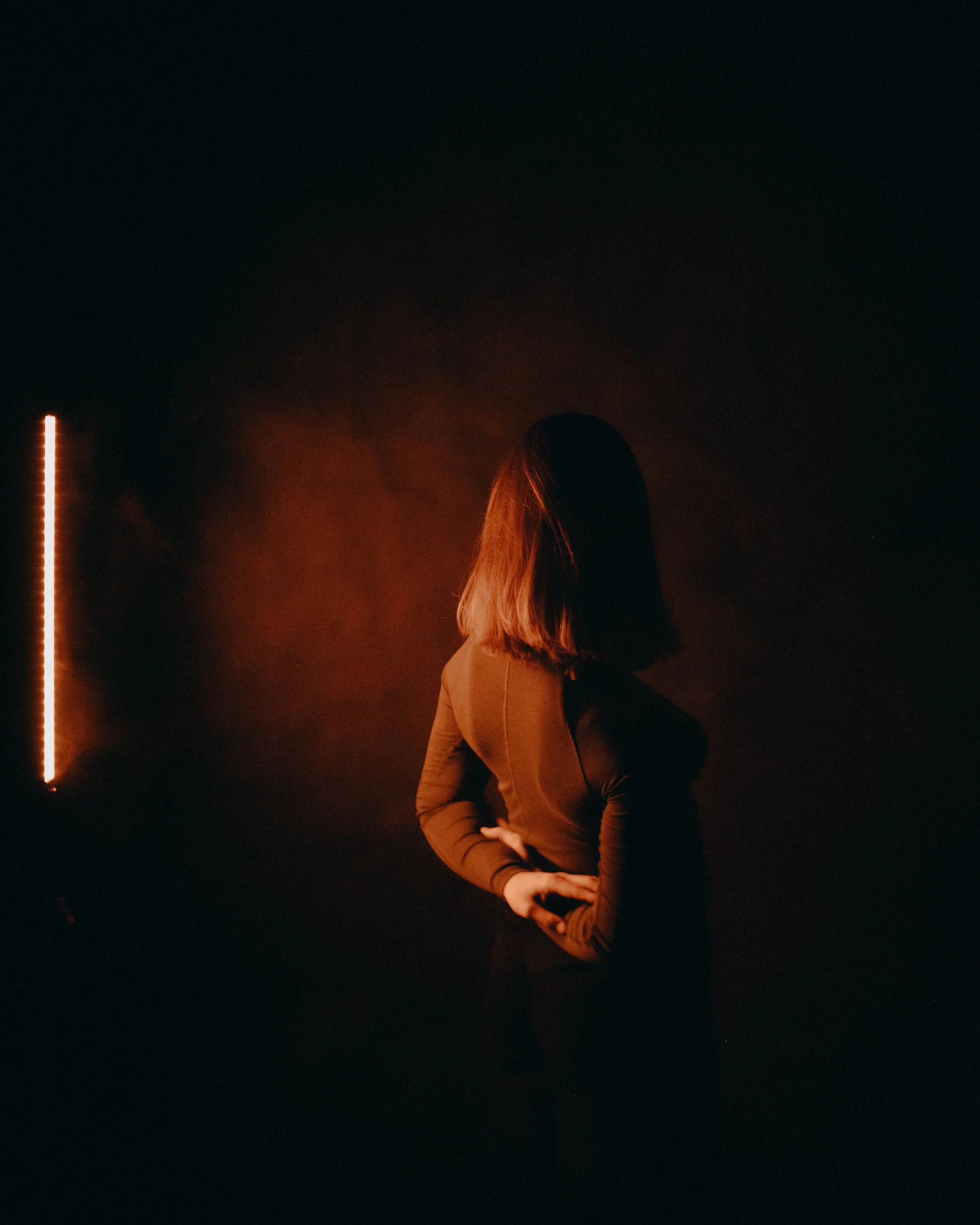 A woman with shoulder-length hair wearing a long-sleeve top, standing in a dark space with a red-orange light on the left side illuminating part of her face and body.