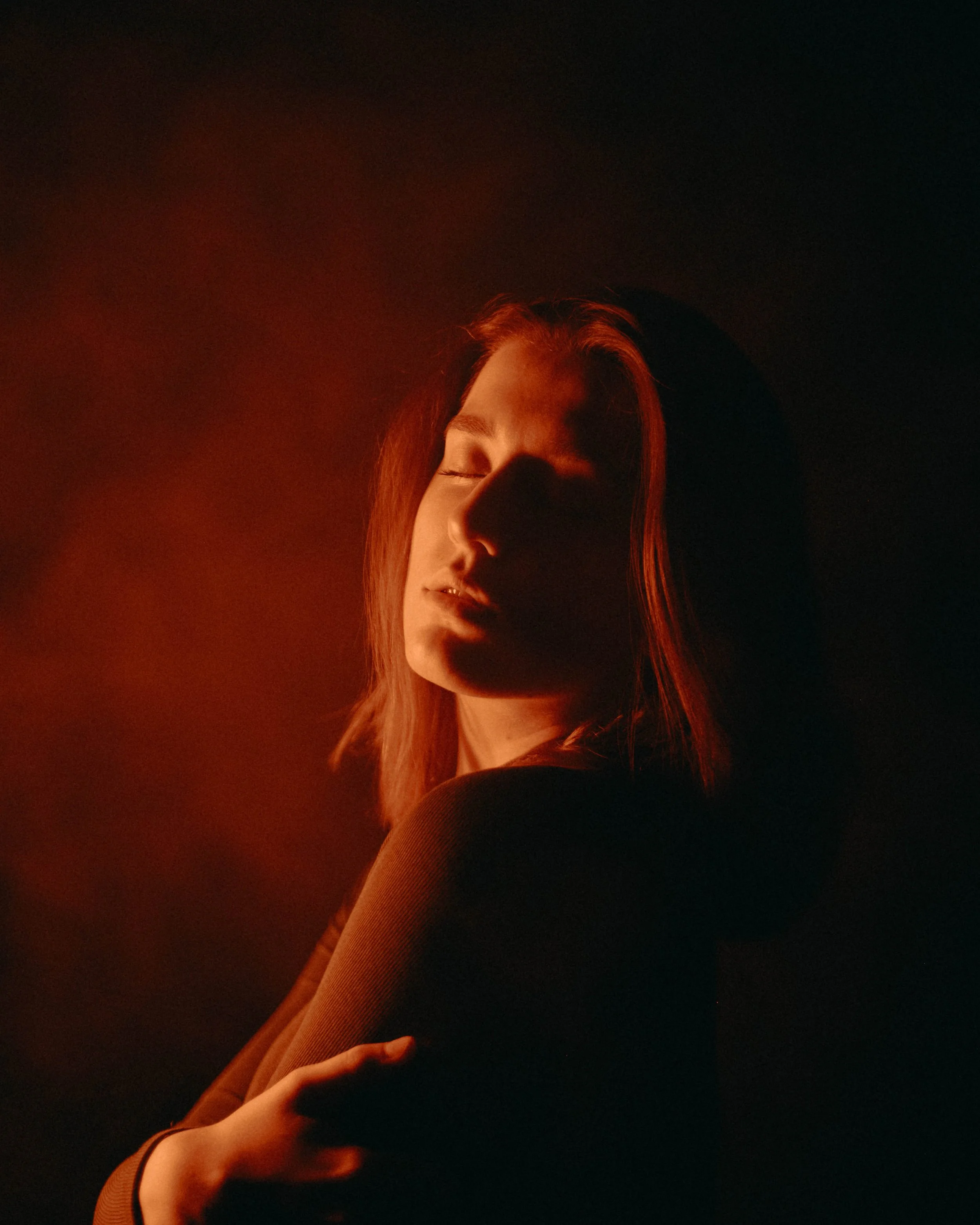 A woman with red hair and closed eyes is illuminated by warm, dramatic lighting against a dark background.