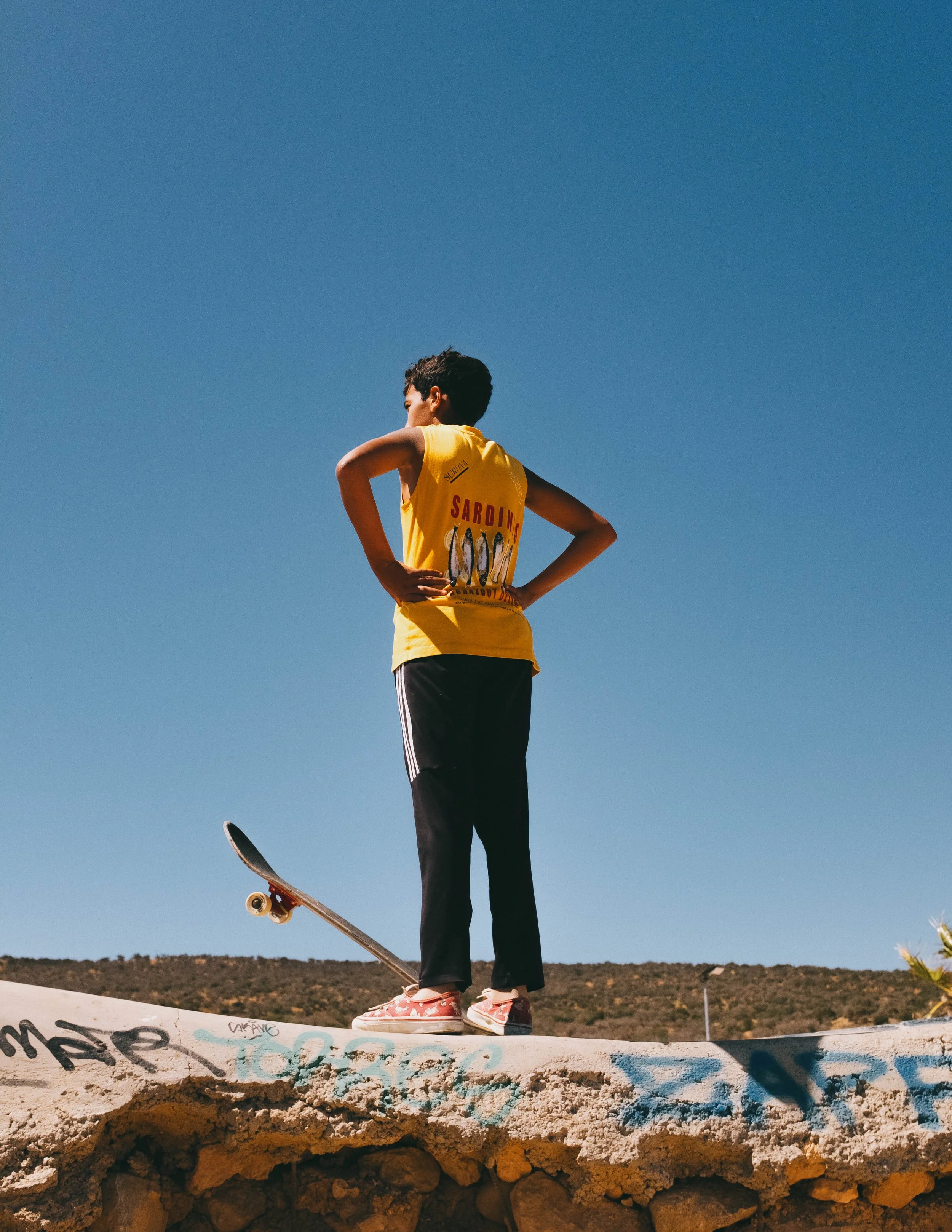 A young boy wearing a yellow T-shirt with a sardine graphic in black and red, black pants with white stripes, and pink shoes, standing on a graffitied concrete ledge with a skateboard under one foot, looking into the distance against a clear blue sky