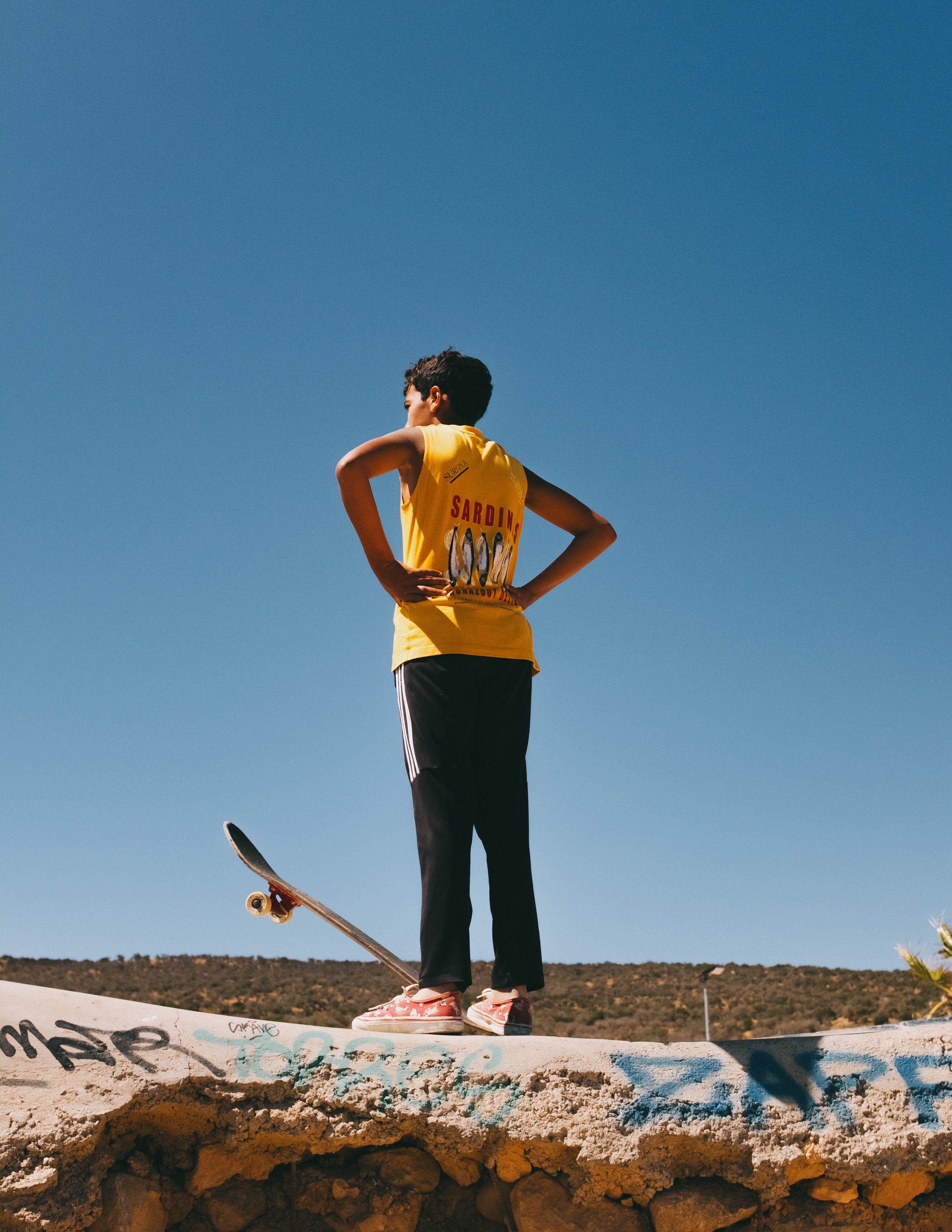 A young boy wearing a yellow T-shirt with a sardine graphic in black and red, black pants with white stripes, and pink shoes, standing on a graffitied concrete ledge with a skateboard under one foot, looking into the distance against a clear blue sky