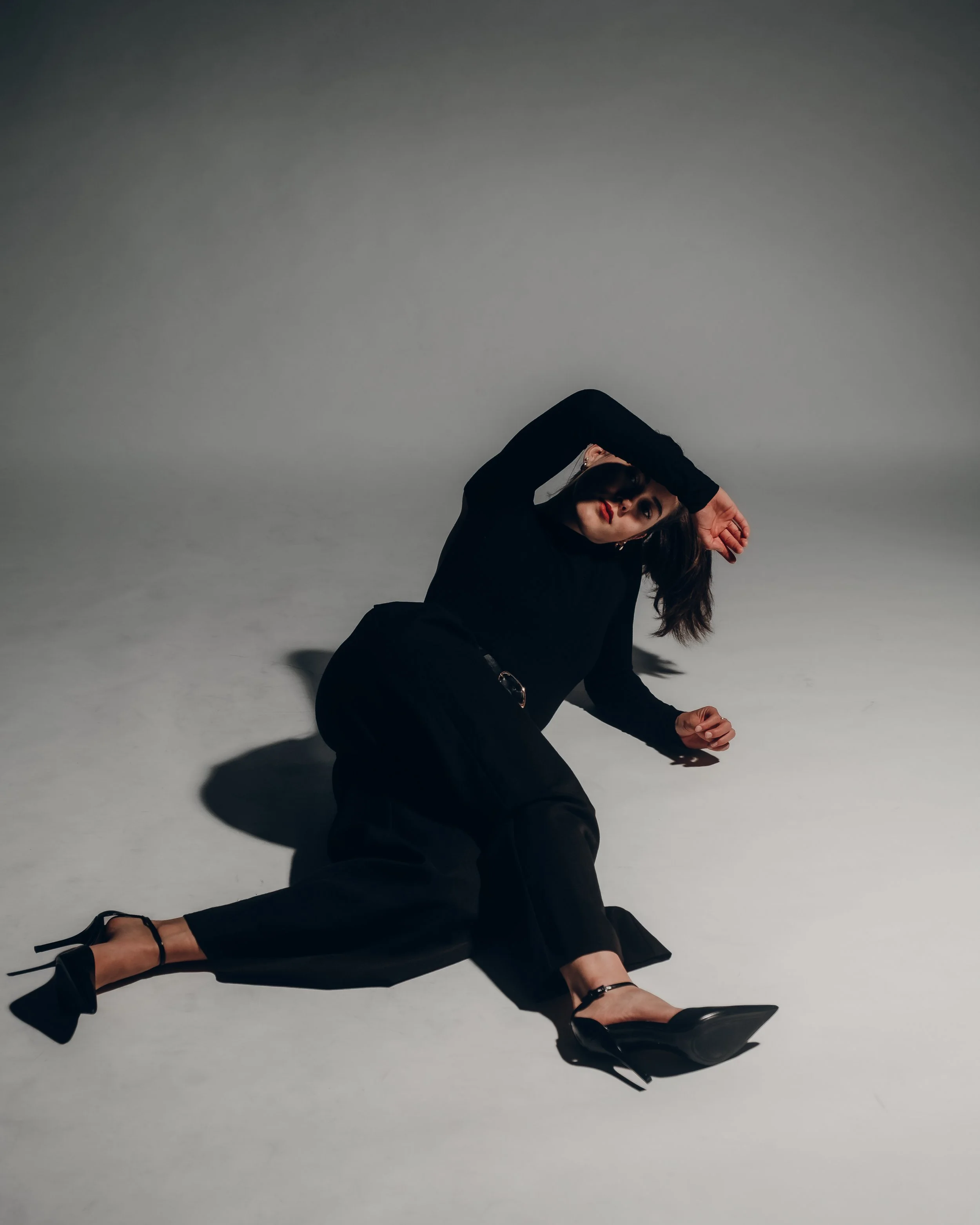 A woman in black clothing is lying on the floor, with her head tilted and one arm covering her eyes, in a studio setting with a plain background.