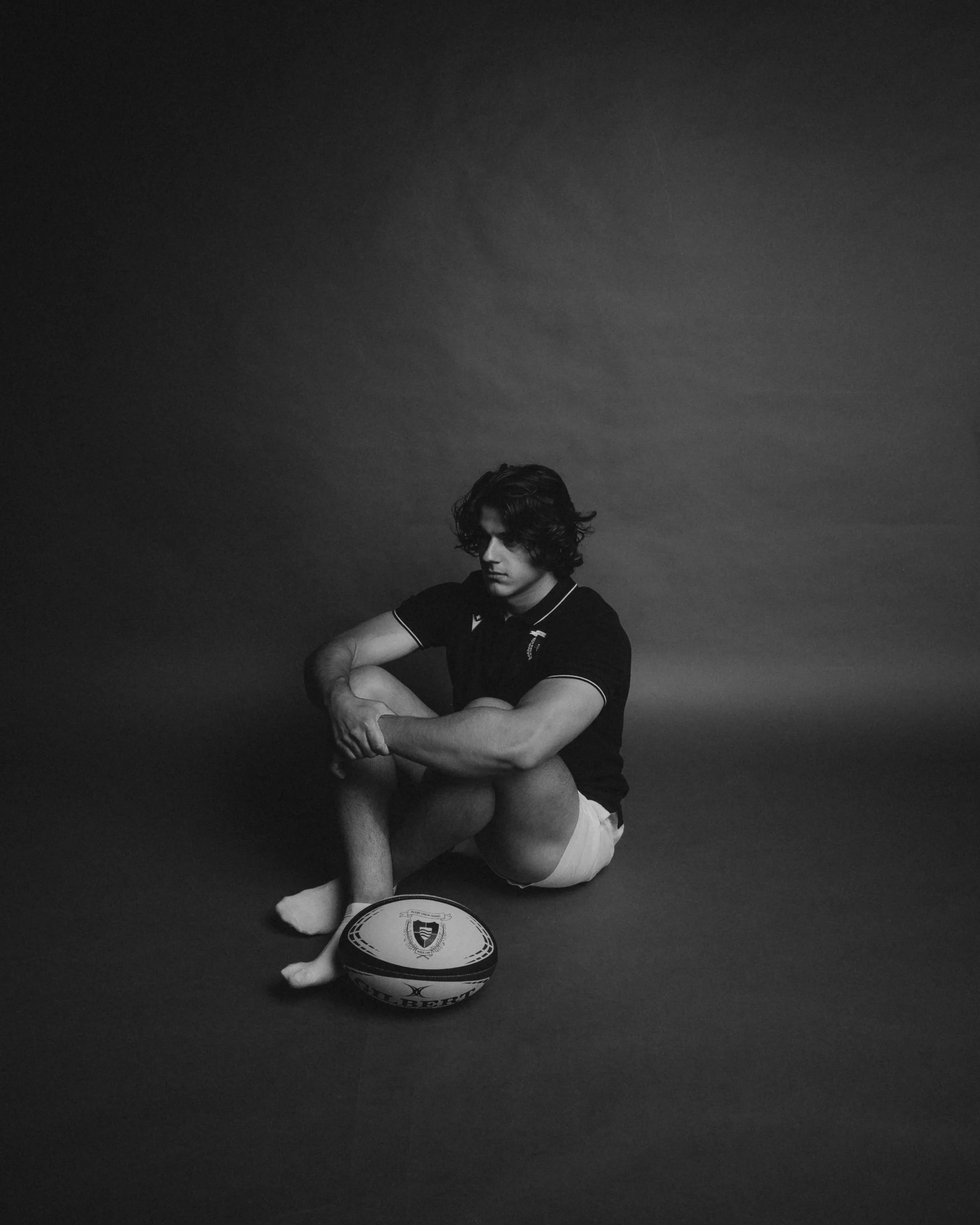 A black and white photo of a young man sitting on the floor with a basketball at his feet. He has wavy hair, is wearing a dark polo shirt and light shorts, and is looking down with a contemplative expression.