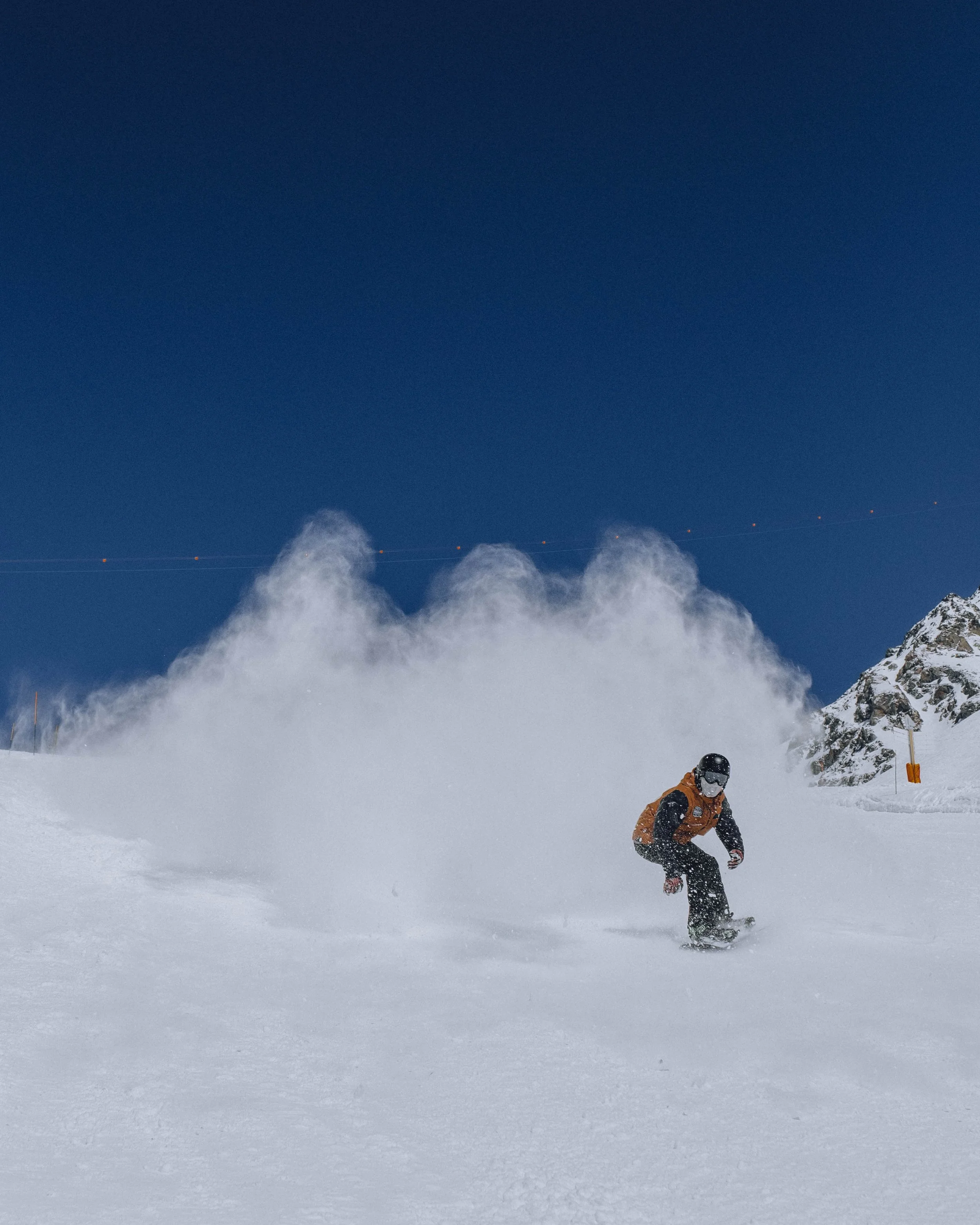 A person snowboarding down a snowy mountain slope, kicking up a spray of snow under a clear blue sky.