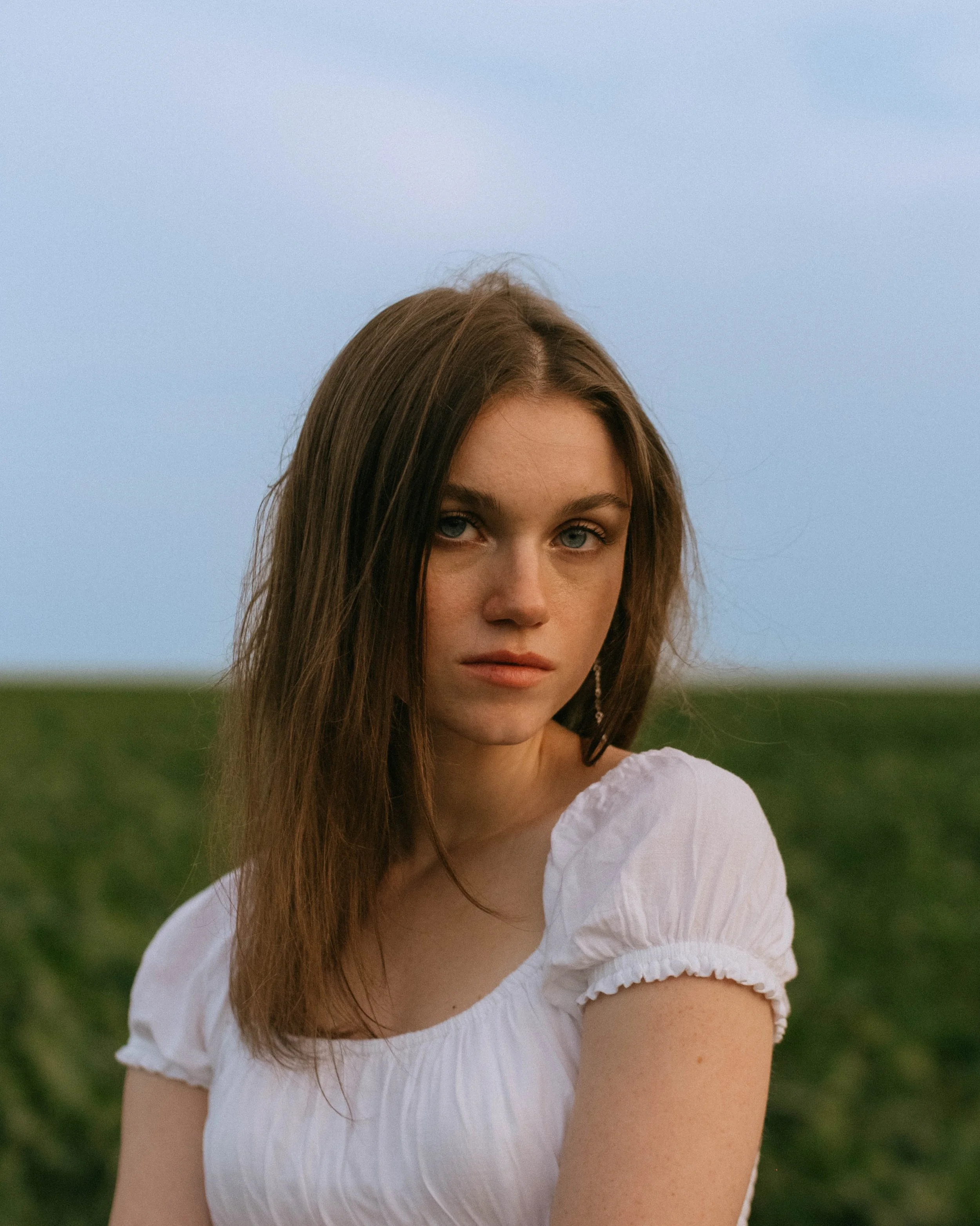 A young woman with long brown hair and blue eyes standing outdoors against a background of green fields and a clear blue sky.