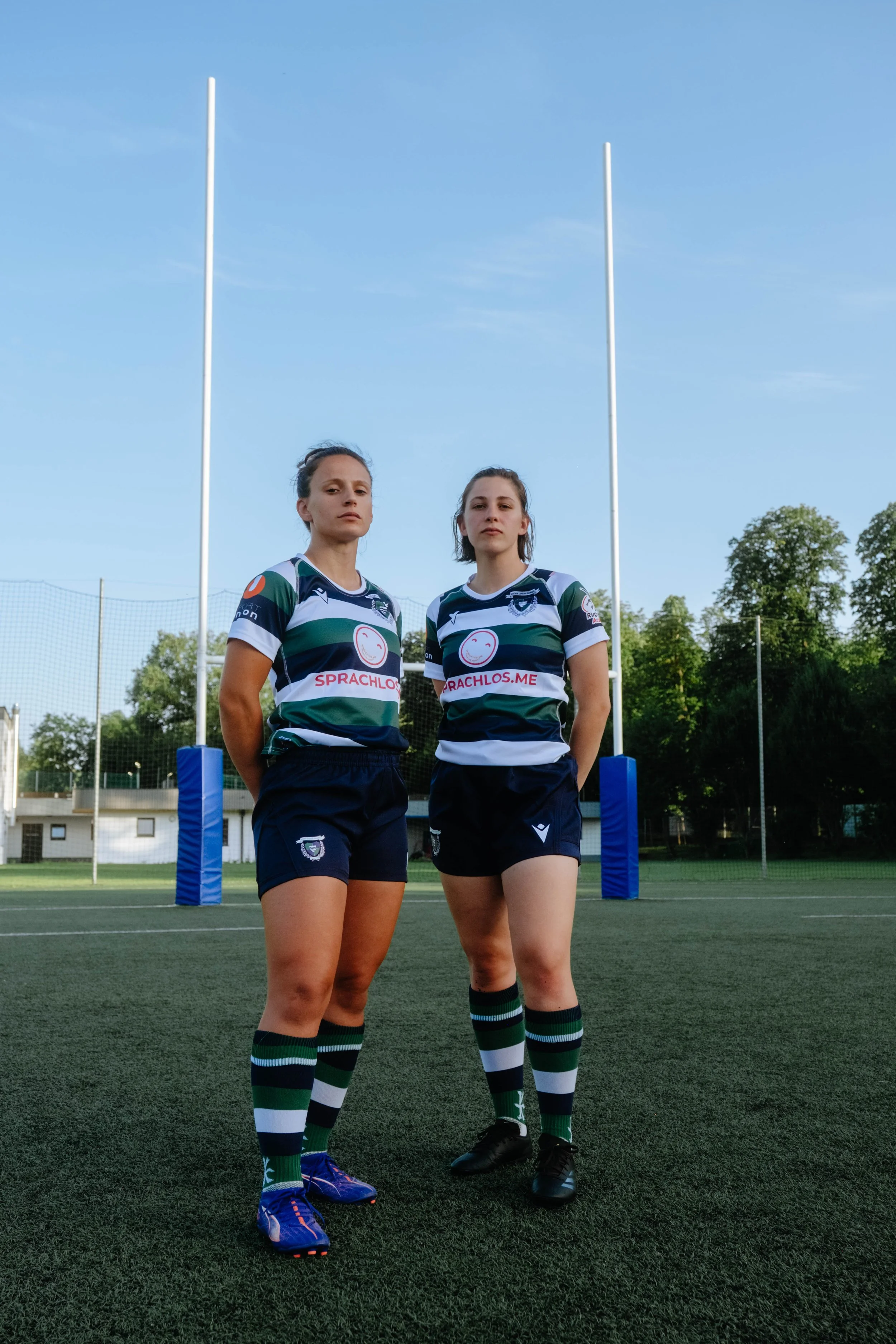 Two female rugby players standing on a rugby field in uniform, with goalposts behind them and trees in the background.