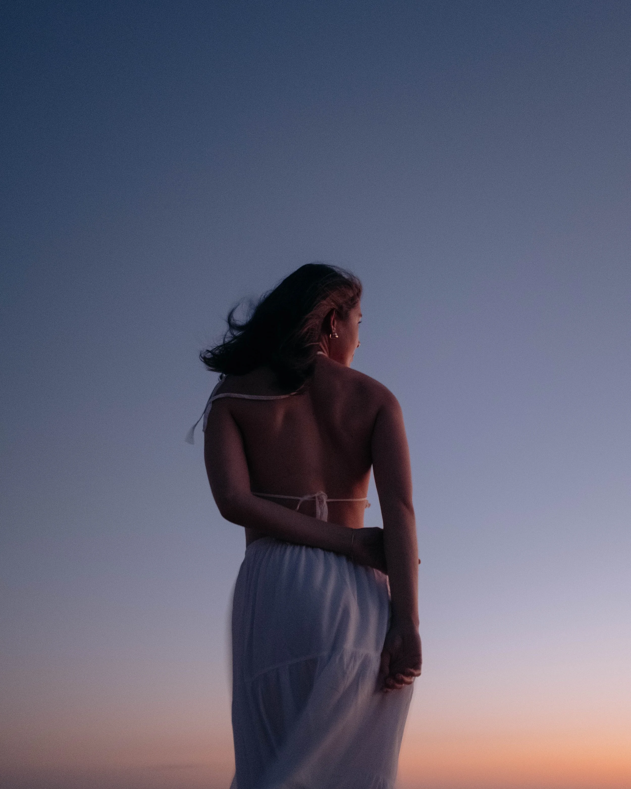 A woman in a white dress with her hair blowing in the wind, standing on a beach at sunset with a clear sky.