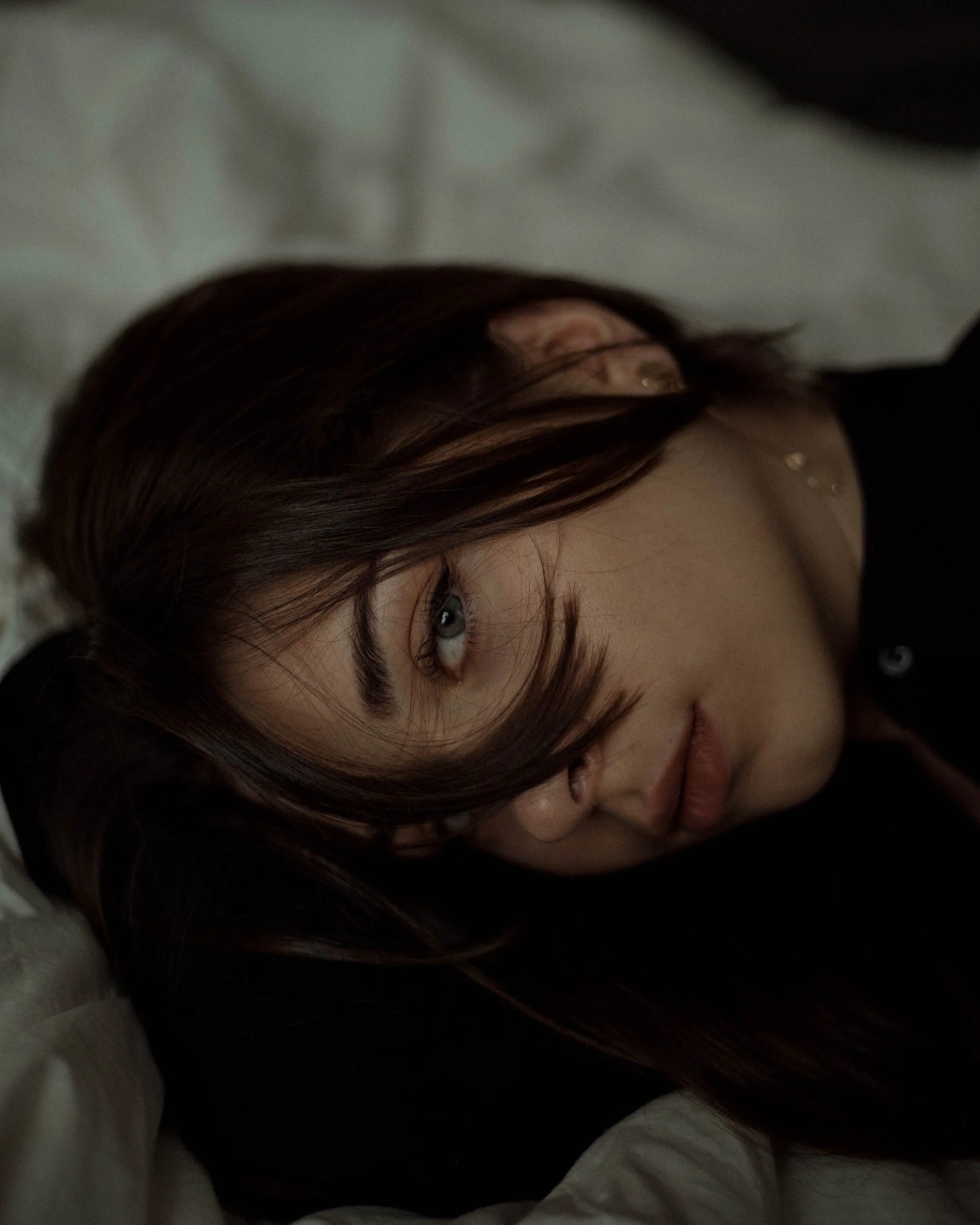 Close-up of a young woman with brown hair, lying down on a white pillow, with her head tilted and looking at the camera.