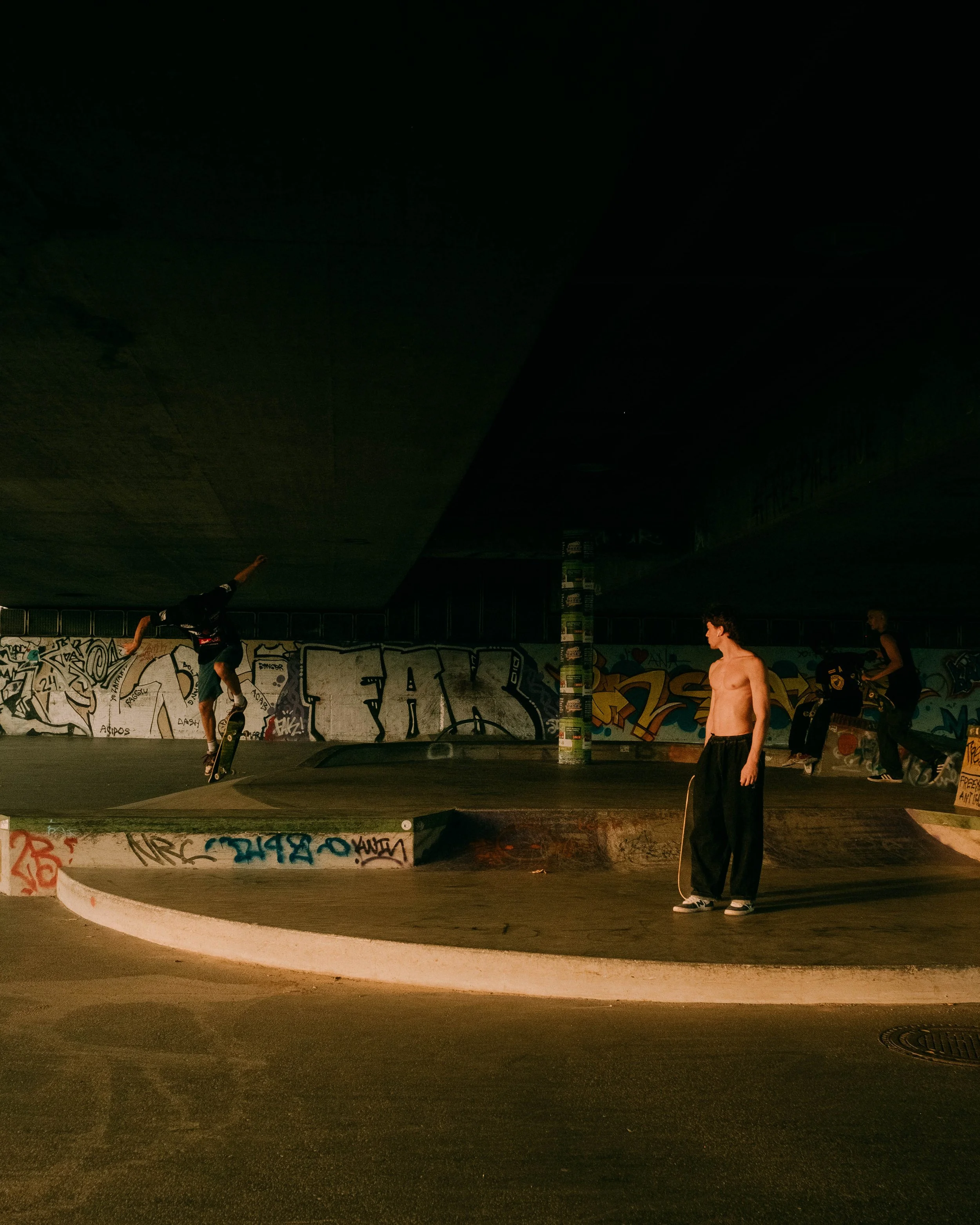 Two skateboarders perform tricks at a skate park under a bridge, with graffiti on the walls and a shirtless young man watching.