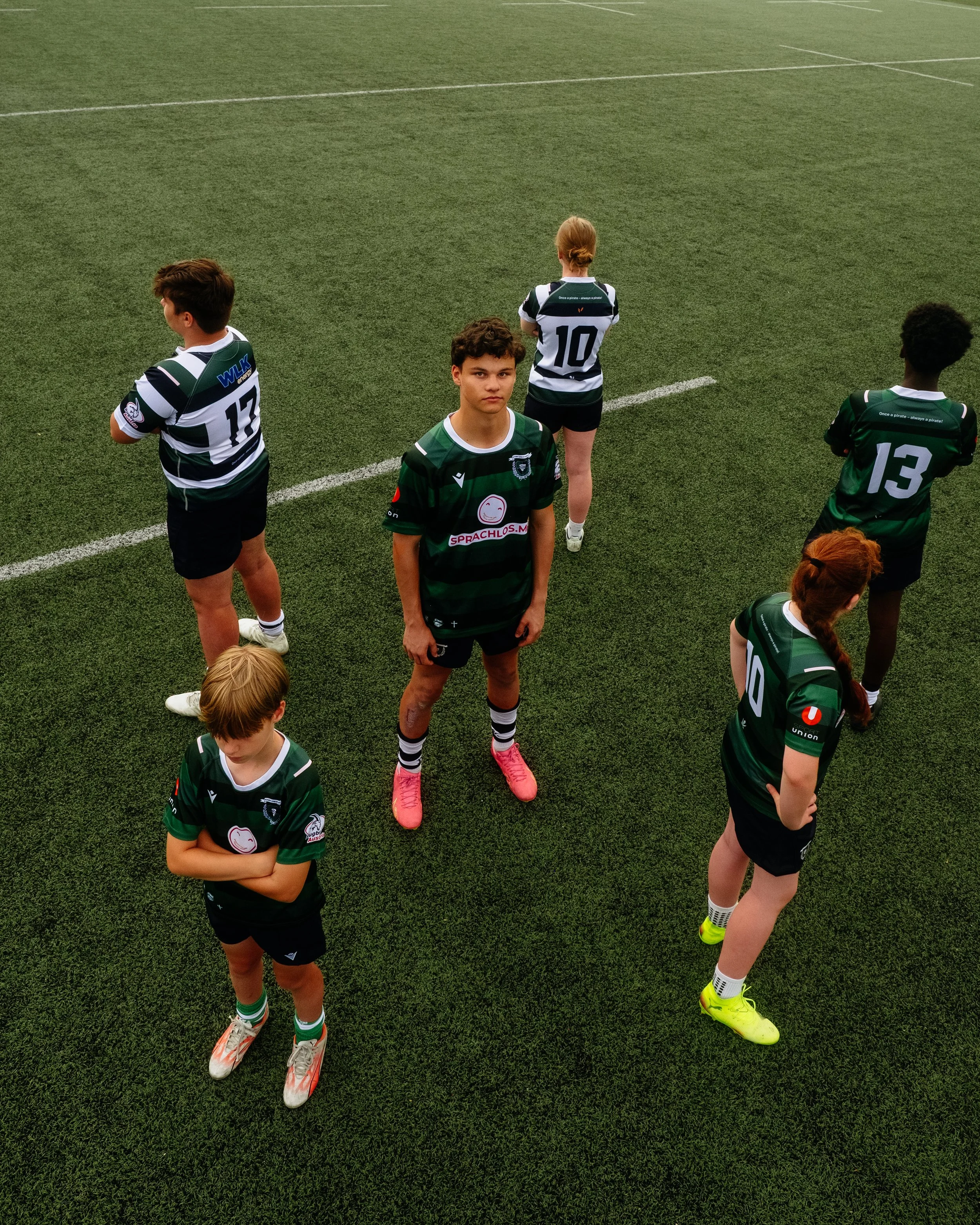 A group of young soccer players on a field, wearing green and black uniforms, with some facing away and one facing forward.