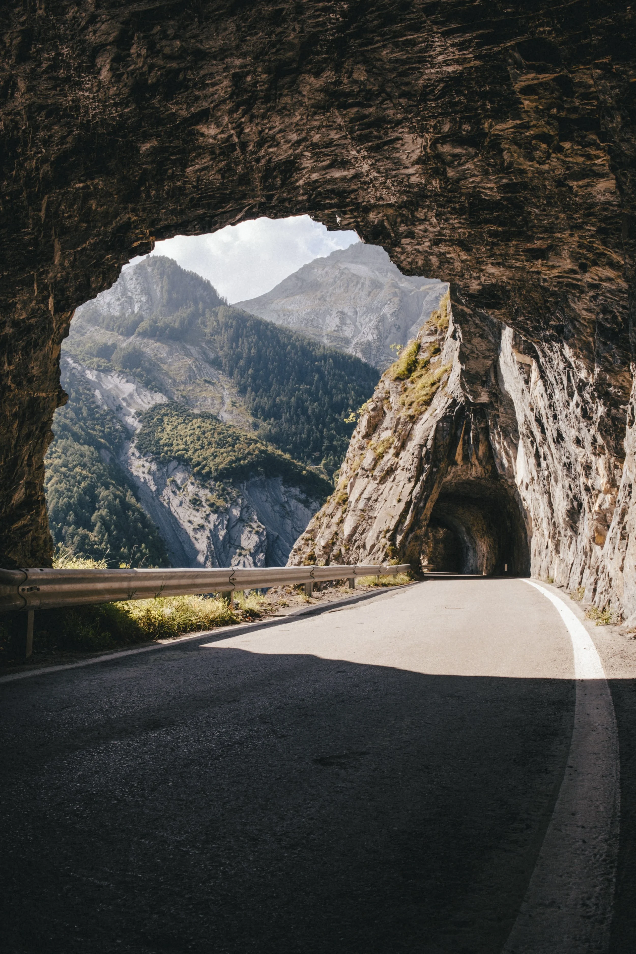 A mountain tunnel with a view of lush green mountains and rocky peaks in the background, sunlight illuminating the tunnel's rocky ceiling and the paved road inside.
