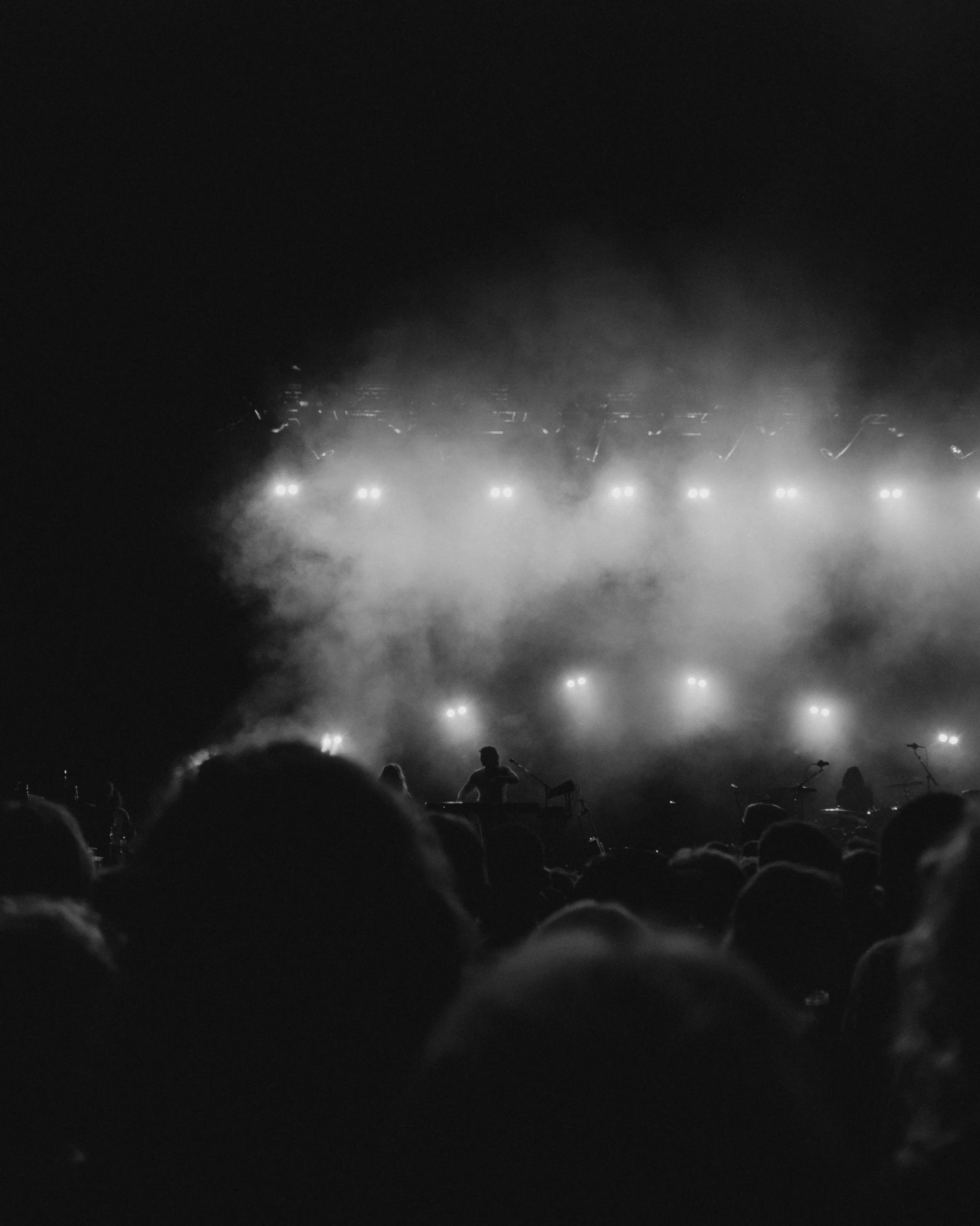 Concert scene with band performing on stage, illuminated by bright lights and surrounded by fog, audience in foreground.