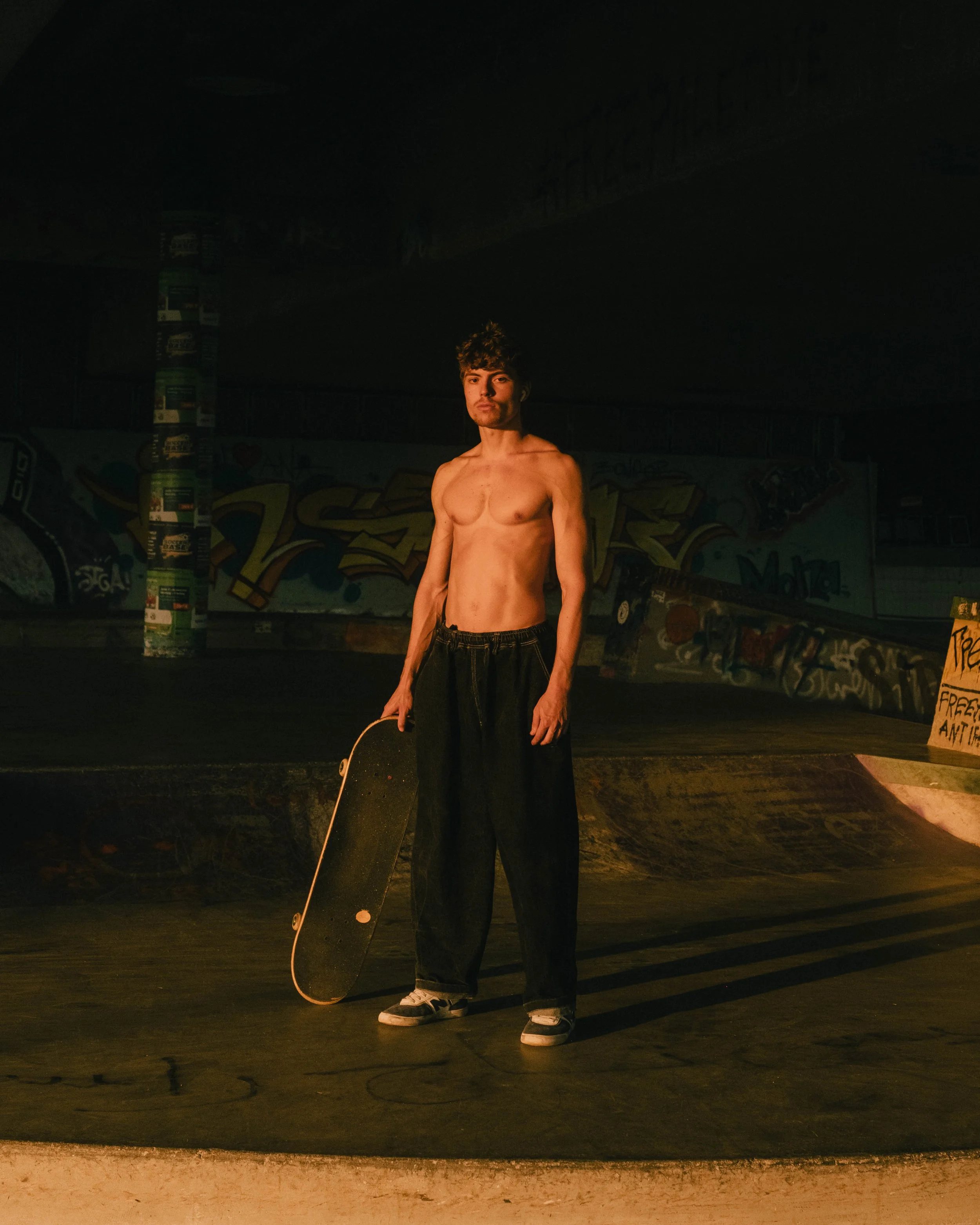 A shirtless young man with dark, curly hair holding a skateboard in a dimly lit skate park with graffiti on the walls.