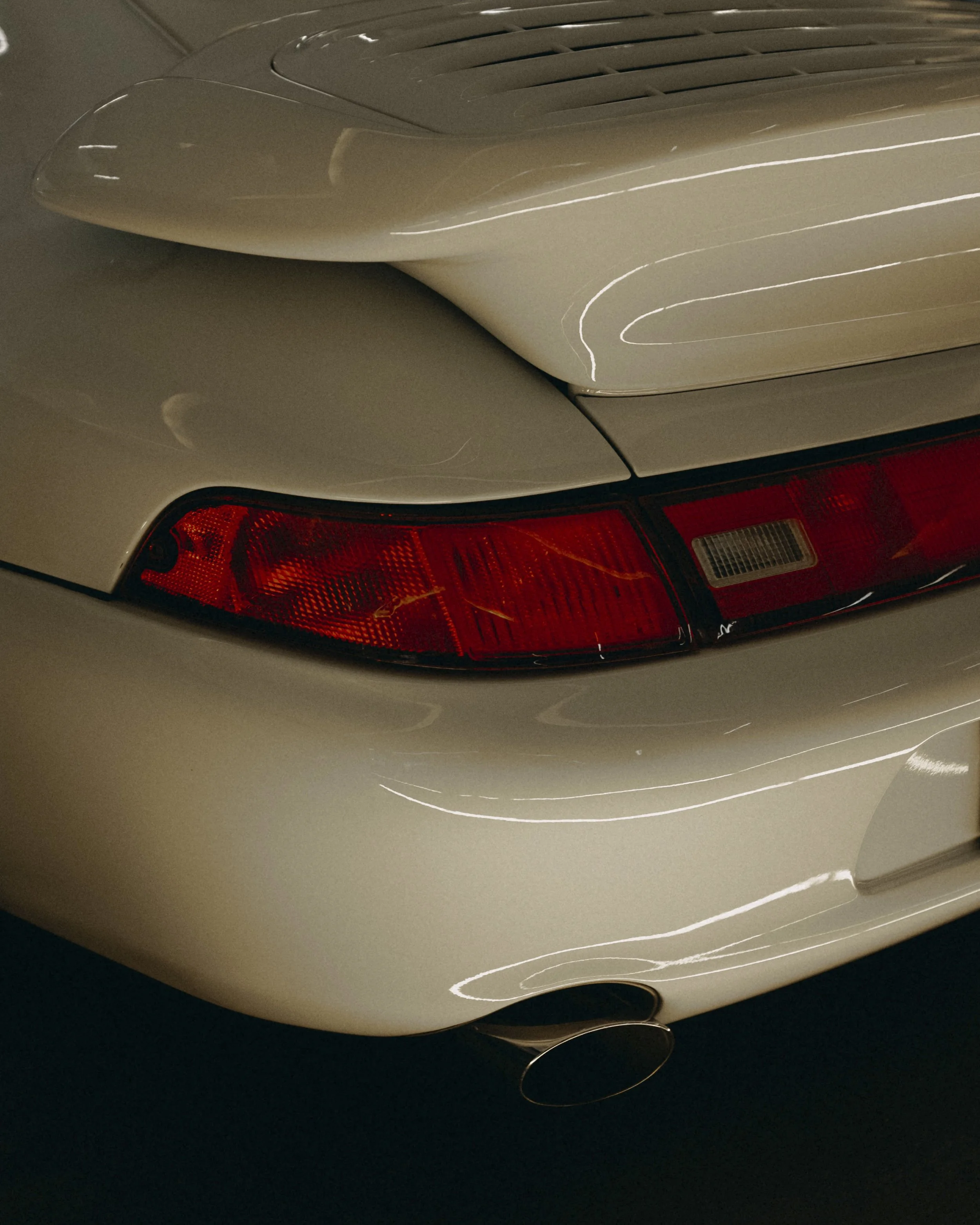 Close-up of the rear end of a beige sports car, showing the tail light, rear spoiler, and exhaust pipe.