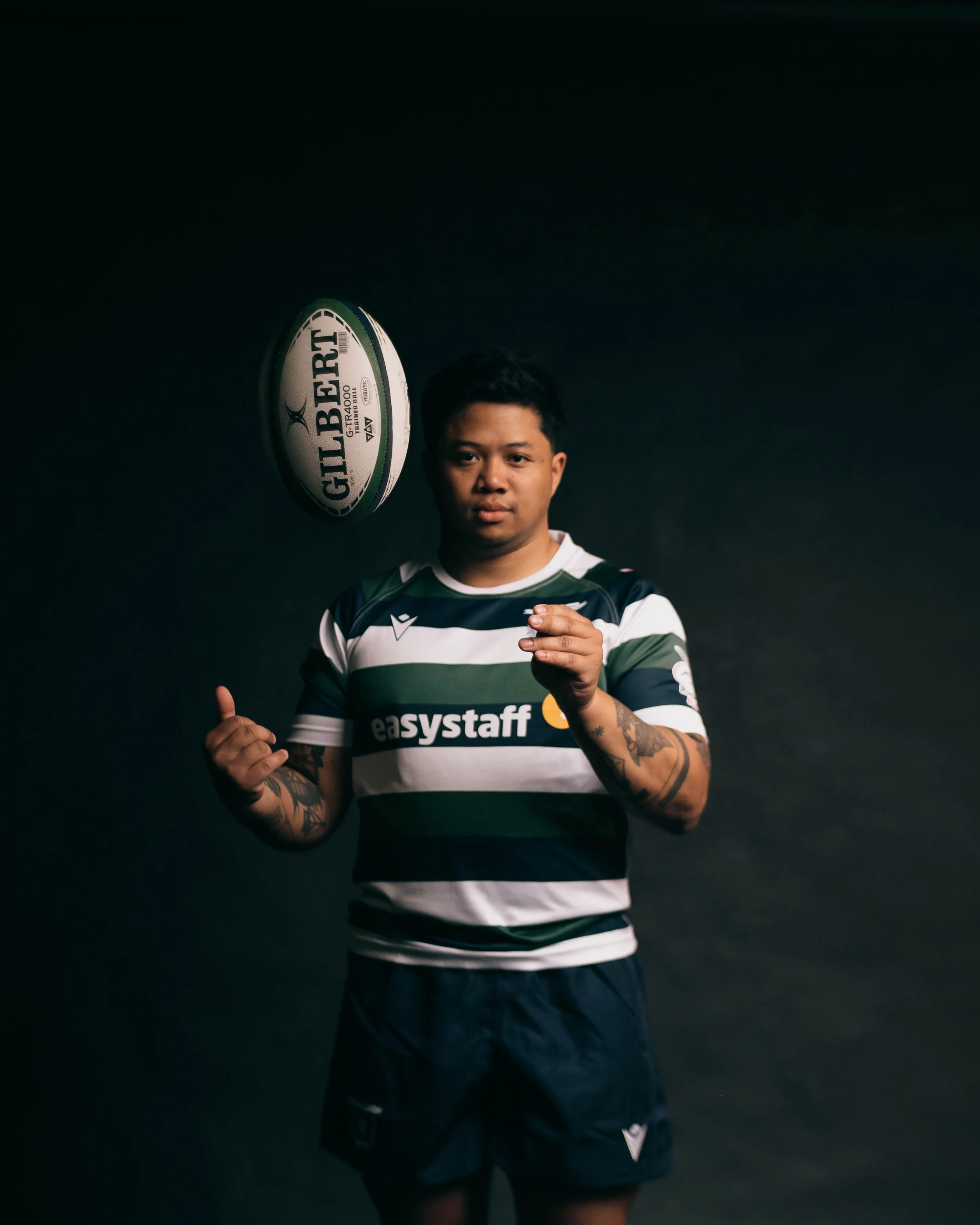 A rugby player in a striped jersey poses with a rugby ball against a black background.