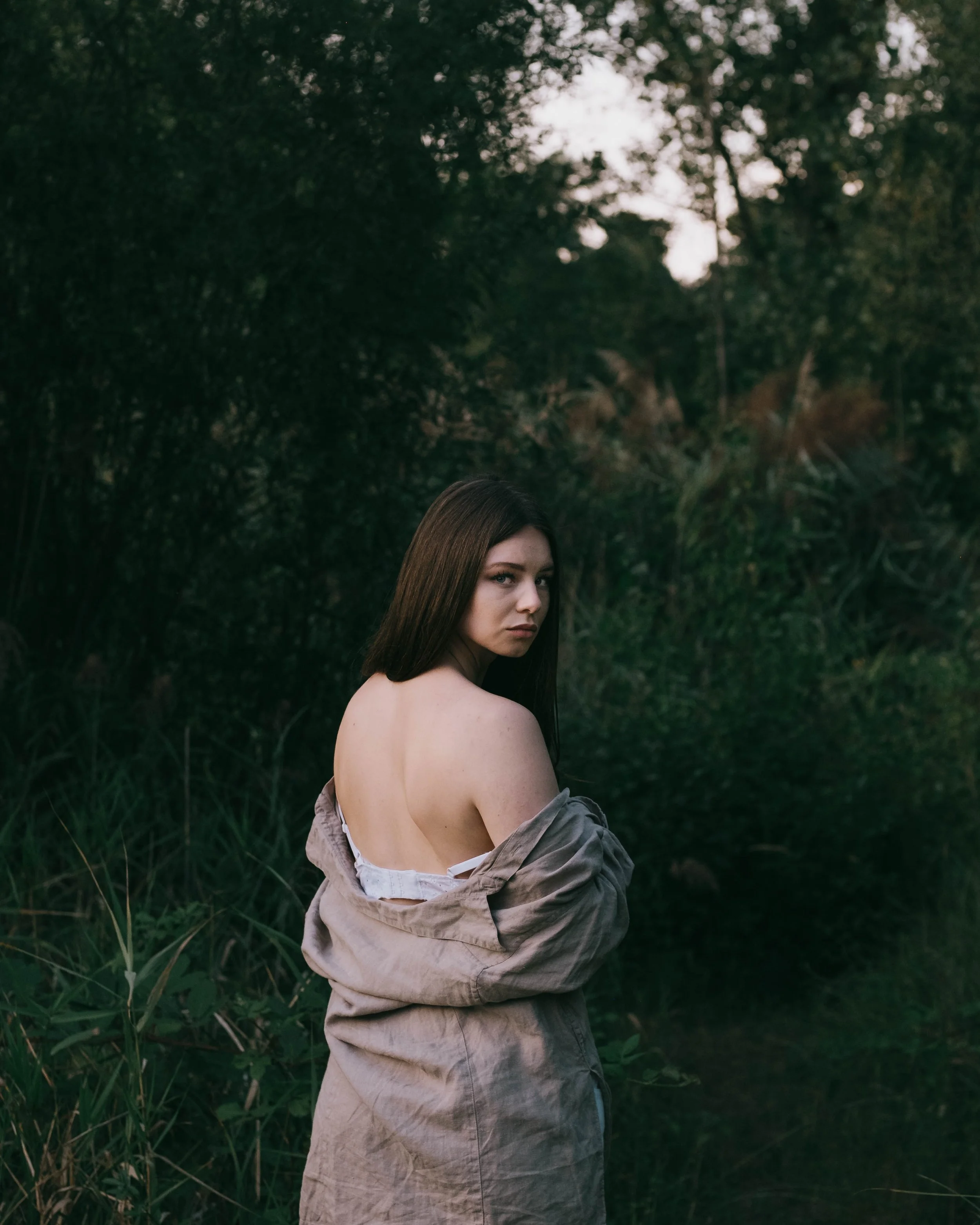 A young woman with long dark hair standing outdoors in a natural setting with green trees and grass, wearing an off-the-shoulder beige jacket and looking over her shoulder at the camera.
