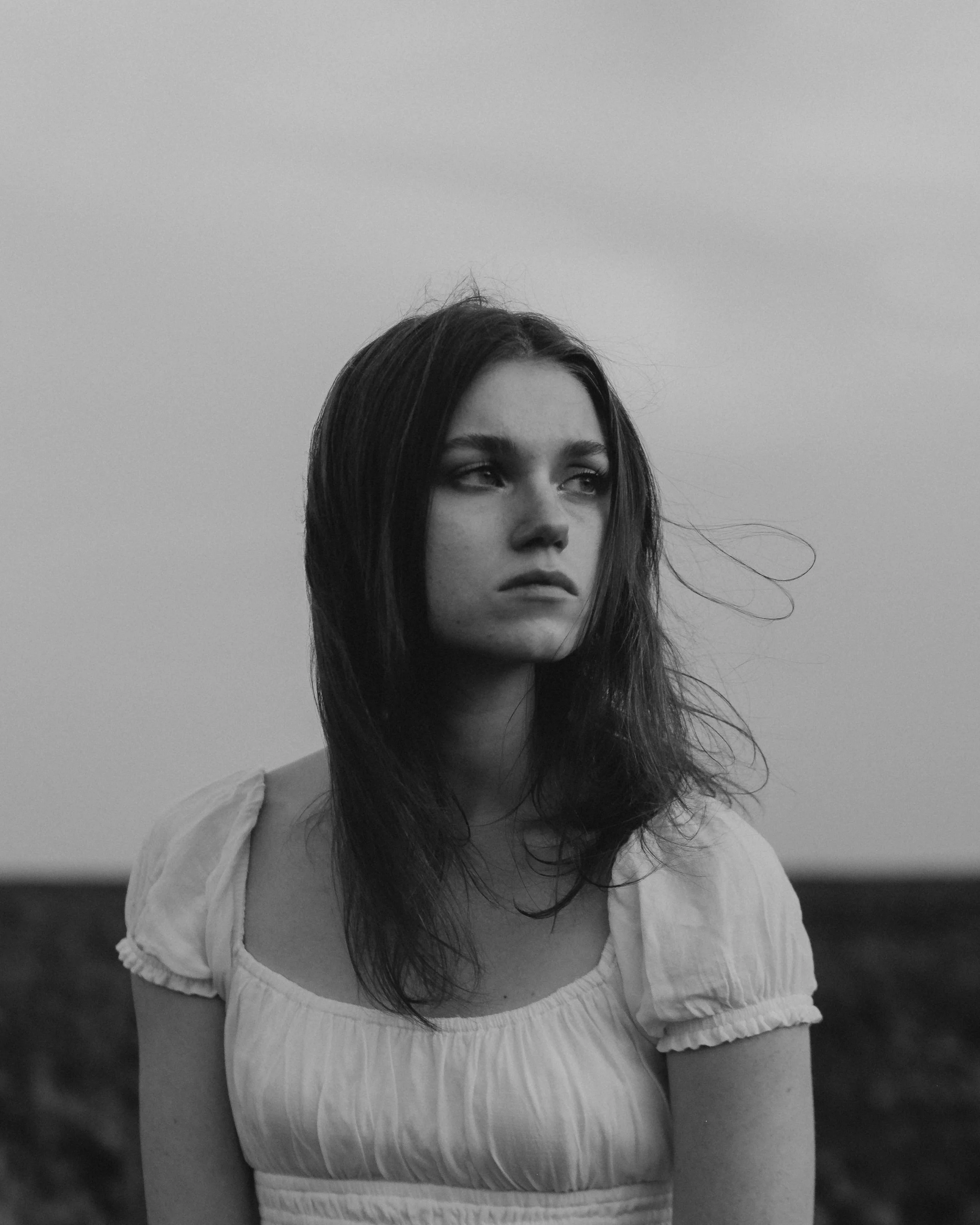 Black and white photo of a young woman with dark hair wearing a light-colored top outdoors, looking to the side with a neutral expression.