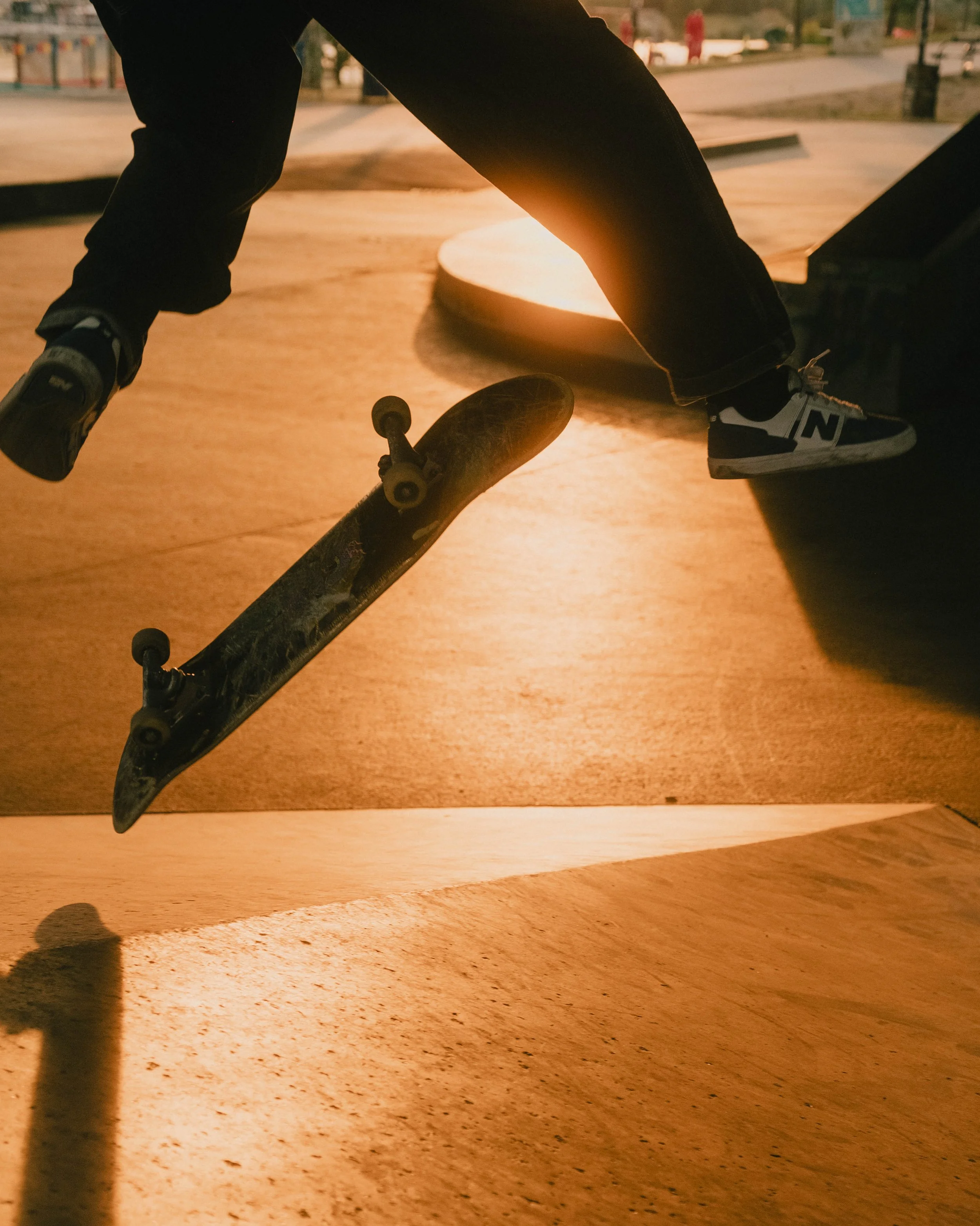 A skateboarder performing a trick in a skate park during sunset, with the skateboard mid-air and the person's legs visible.