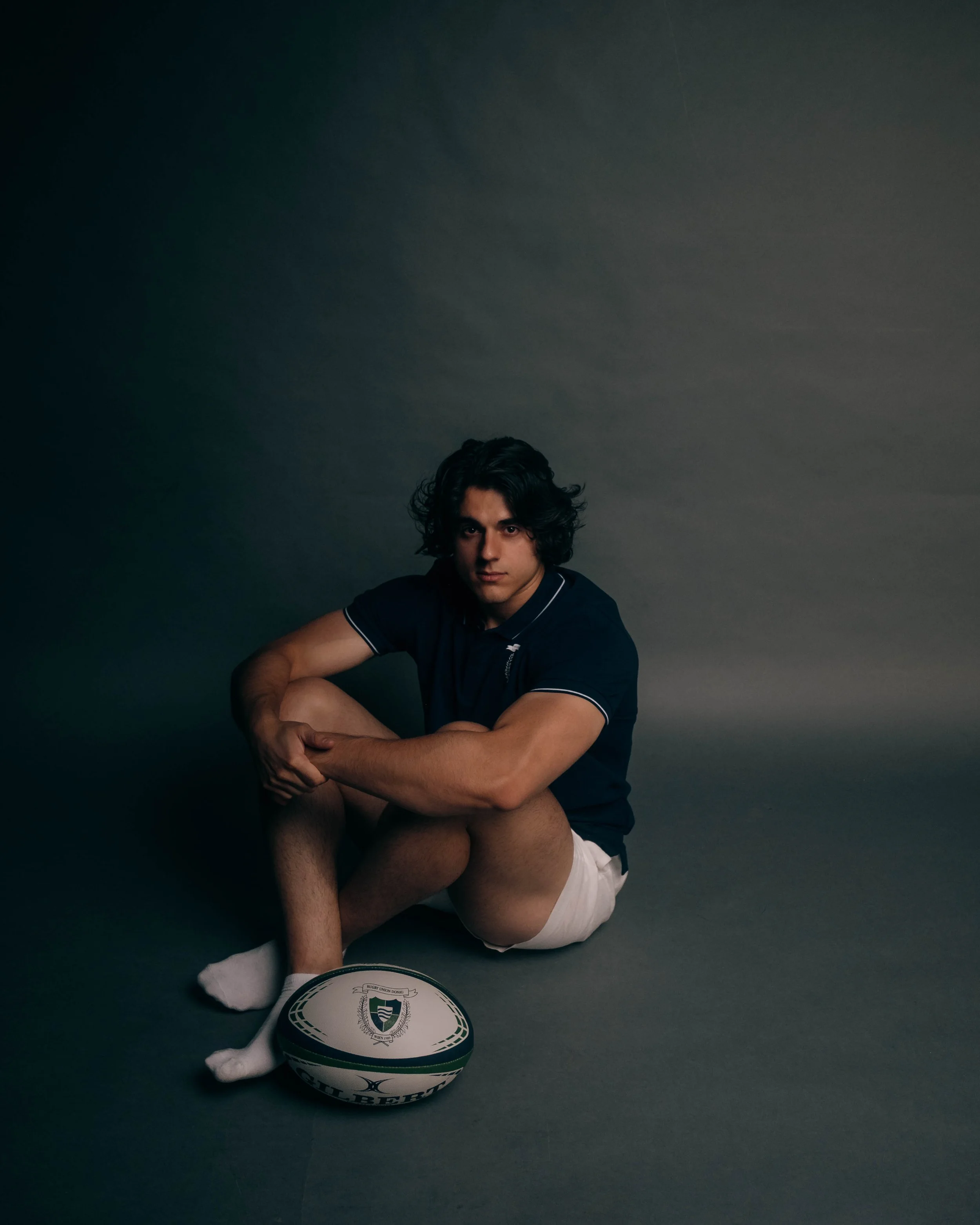 A young man with dark, curly hair sits on the floor wearing a navy blue polo shirt, white shorts, and white socks, with a rugby ball in front of him against a dark, neutral background.